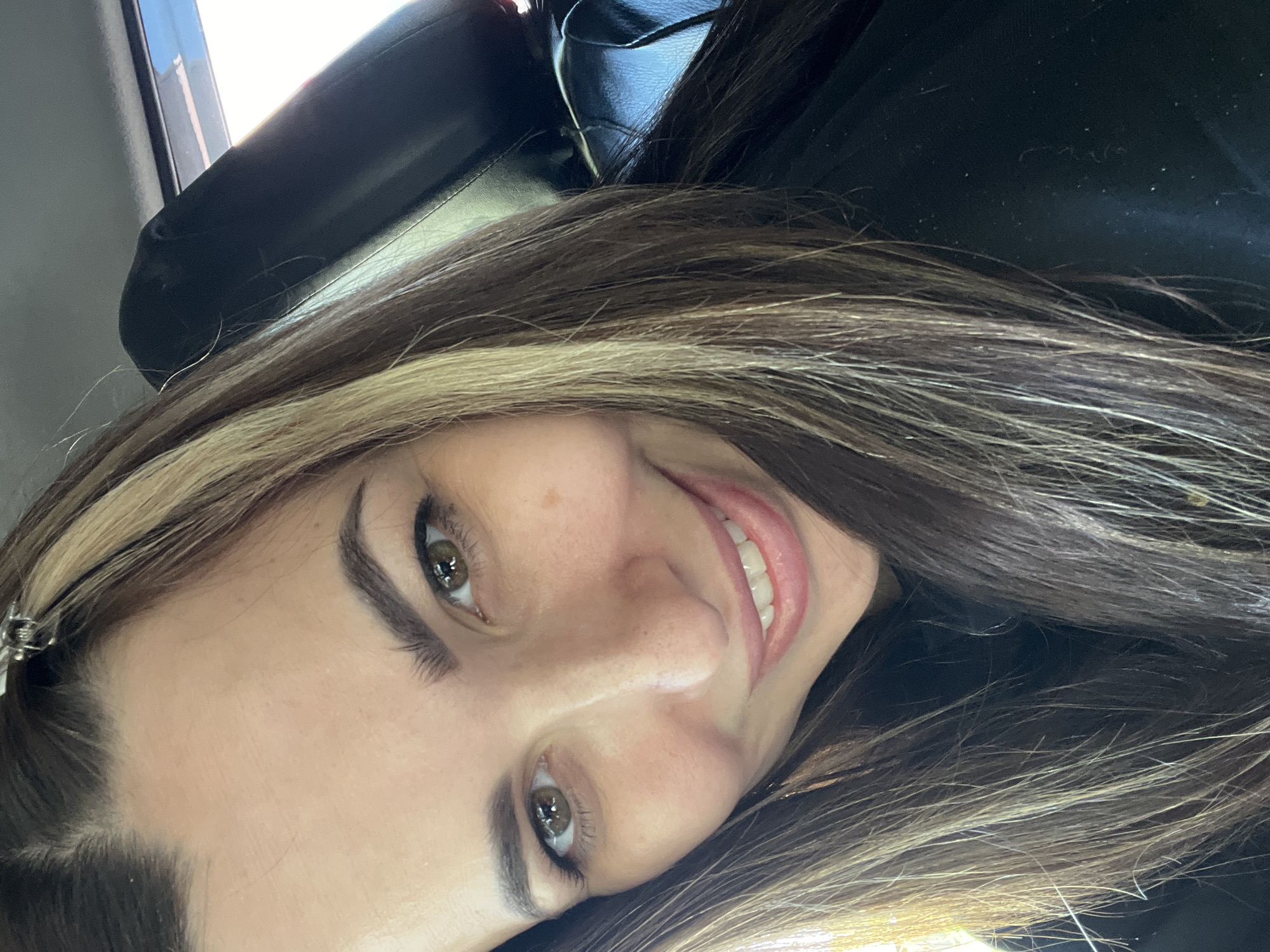 A close-up portrait shows a woman with long, brown hair and blonde highlights smiling at the camera. She has light skin, defined eyebrows, and appears to be wearing eyeliner. The background includes a black leather seat and part of a window. The location is unknown.