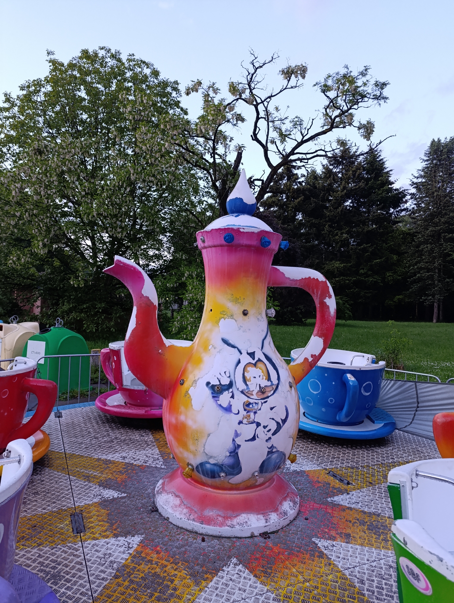 The image shows a colorful teapot-themed ride in what appears to be an amusement park or a fairground. The teapot is predominantly white, orange, and pink, with cartoonish characters painted on its surface. Several teacup-shaped ride cars surround the teapot. The ground is a patterned metal platform. In the background, there are green trees and a clear sky. The location is in the city of Sofia, Bulgaria.