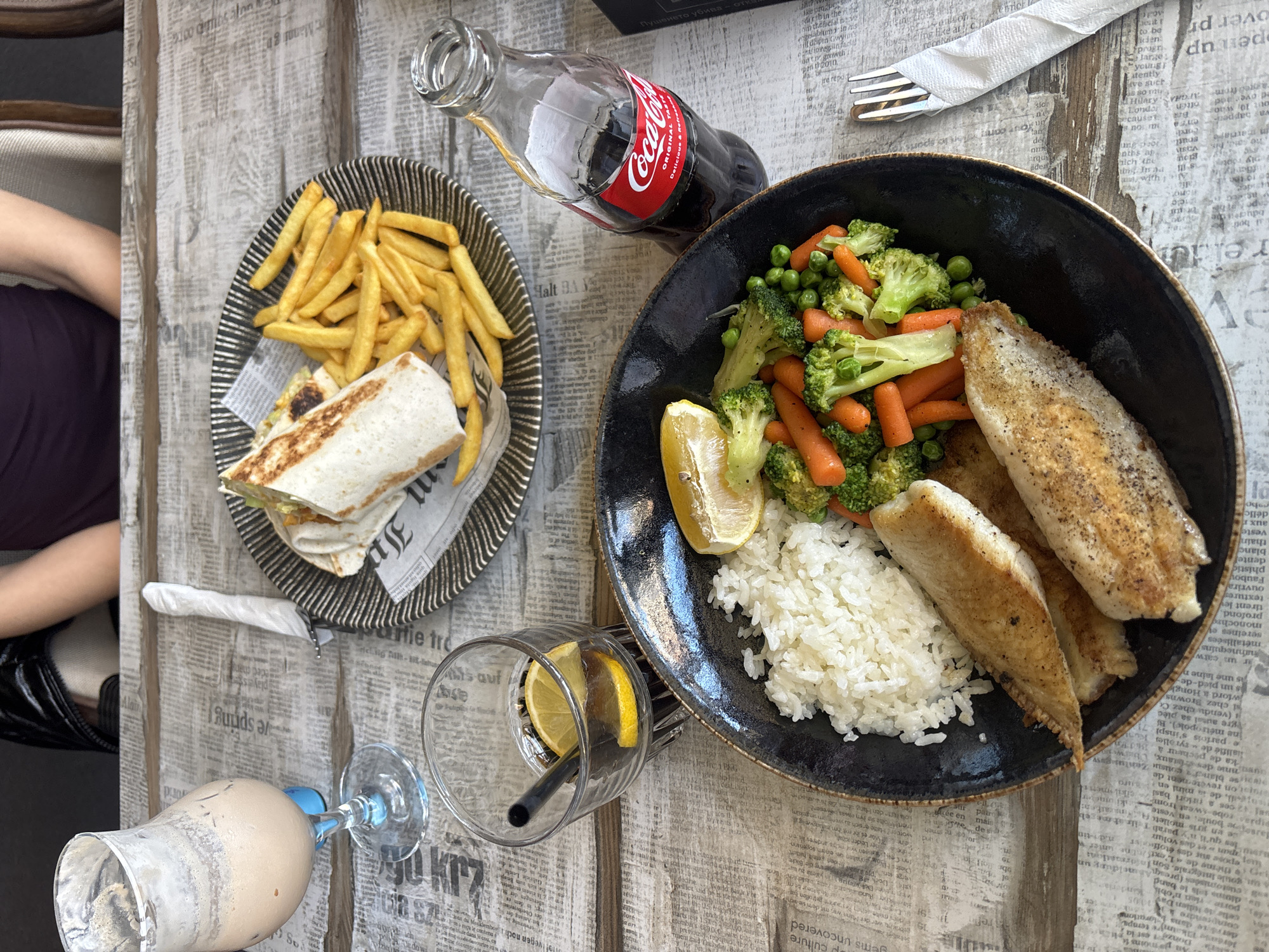 **Scene:** A table in Plovdiv, Bulgaria, set for a meal.
**Subjects:** A portion of a person is visible, wearing a dark purple garment and seated in a tan chair.
**Objects:**
* A black plate containing fried fish, white rice, a lemon wedge, broccoli, carrots, and peas.
* A plate with a wrap and french fries.
* A glass with lemon slices and a straw.
* A Coca-Cola bottle.
* A tall glass with a milkshake.
* A napkin and fork.
* The table surface has a newspaper print.
**Environment:** Outdoor setting, likely a restaurant or cafe. The table covering suggests a themed setting.