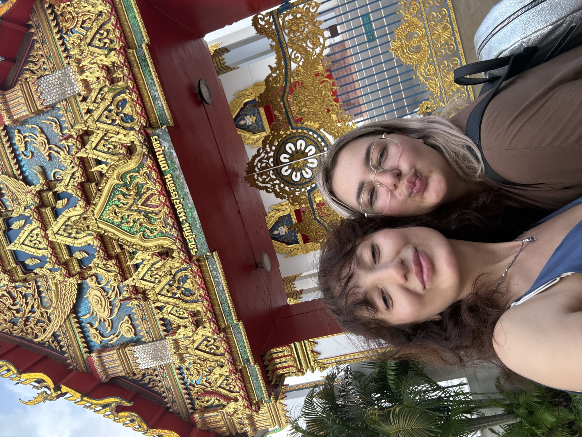 Two young women are taking a selfie in front of a Thai temple entrance. The woman on the left has dark, wavy hair and is wearing a blue dress. The woman on the right has blonde hair and is wearing glasses and a brown top. She is puckering her lips. Behind them, the temple features ornate golden decorations and intricate carvings on a red background. Palm trees are visible to the left of the entrance. The temple is likely located in Phuket City Municipality, Thailand, given the style of architecture.