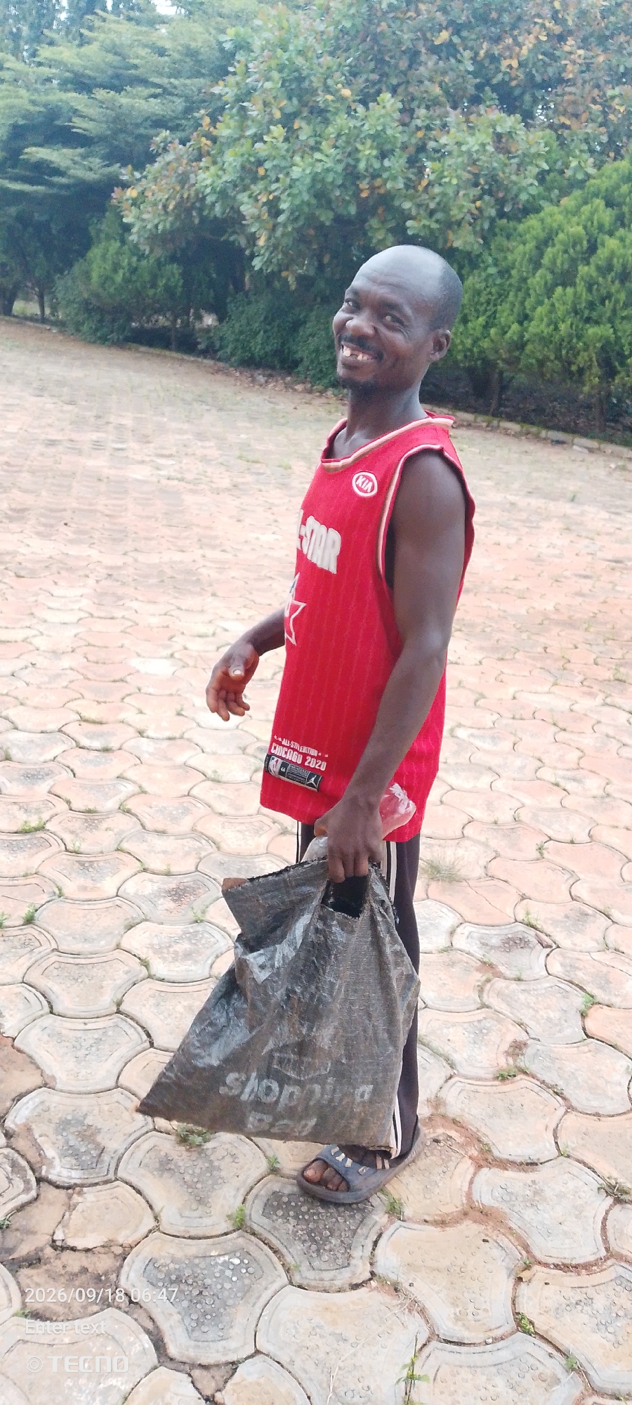 An African man stands on a paved walkway, smiling. He is wearing a red basketball jersey with "STAR" and "CHICAGO 2020" written on it, and black pants. He holds a large black plastic bag in his left hand, and a clear plastic bottle in his right hand. The background shows trees and bushes. The walkway is made of interlocking concrete pavers. The timestamp "2026/09/18 06:47" and "TECNO" are watermarked on the bottom left of the image.