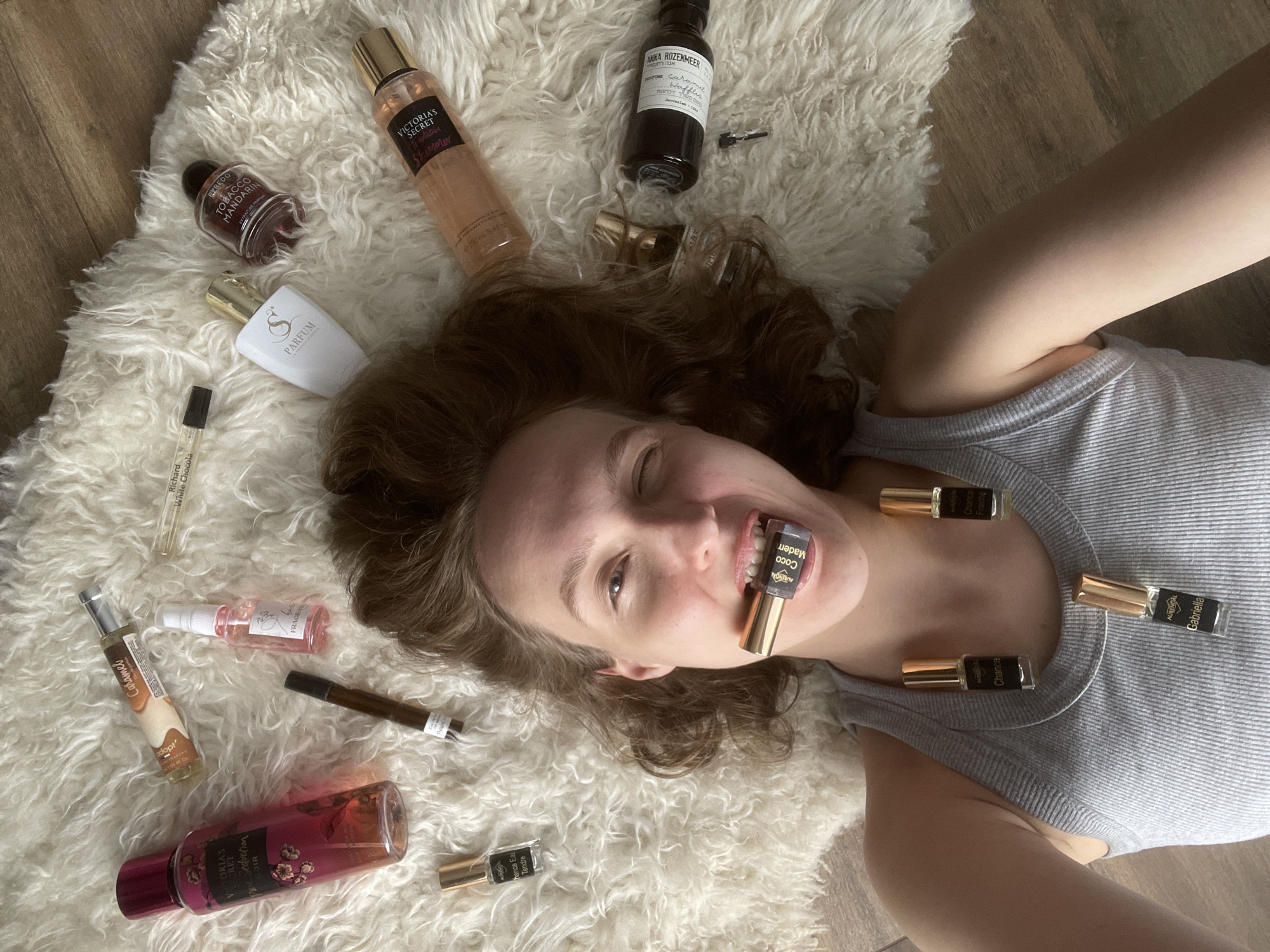 A woman with light skin and brown hair lies on a white rug atop a wooden floor. She wears a gray tank top and has multiple small perfume bottles positioned around her face and on her upper chest. One bottle is held in her mouth. Various perfume bottles and sprays are scattered around her on the rug, including brands like Victoria's Secret, Byredo, Anna Rozenmeer, and Adopt. Some of the perfumes are labeled "Caramel" and "Coco Mademoiselle." The location isn't identified as Podshivalovo, Russia in the image.