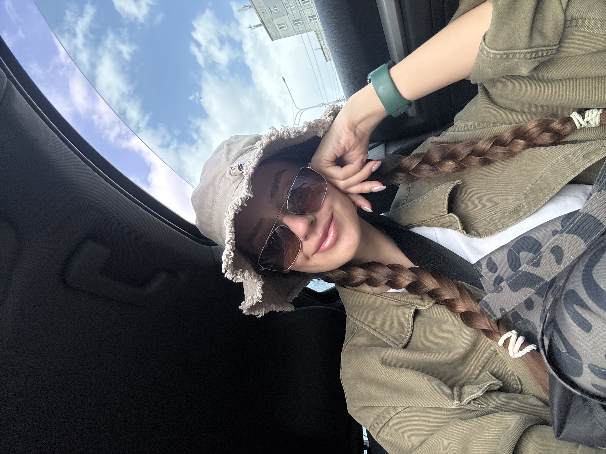 A young woman is taking a selfie from inside a car. She is wearing a light brown bucket hat with a frayed edge and sunglasses with brown lenses. She has long brown braids that are secured with white yarn. She is wearing a light brown jacket and a green watch on her left wrist. The car has a sunroof through which a blue sky with white clouds is visible. Outside the car, there is a tall apartment building and a streetlight. Based on the architecture, the location is likely in the city of Krasnoyarsk, Russia.