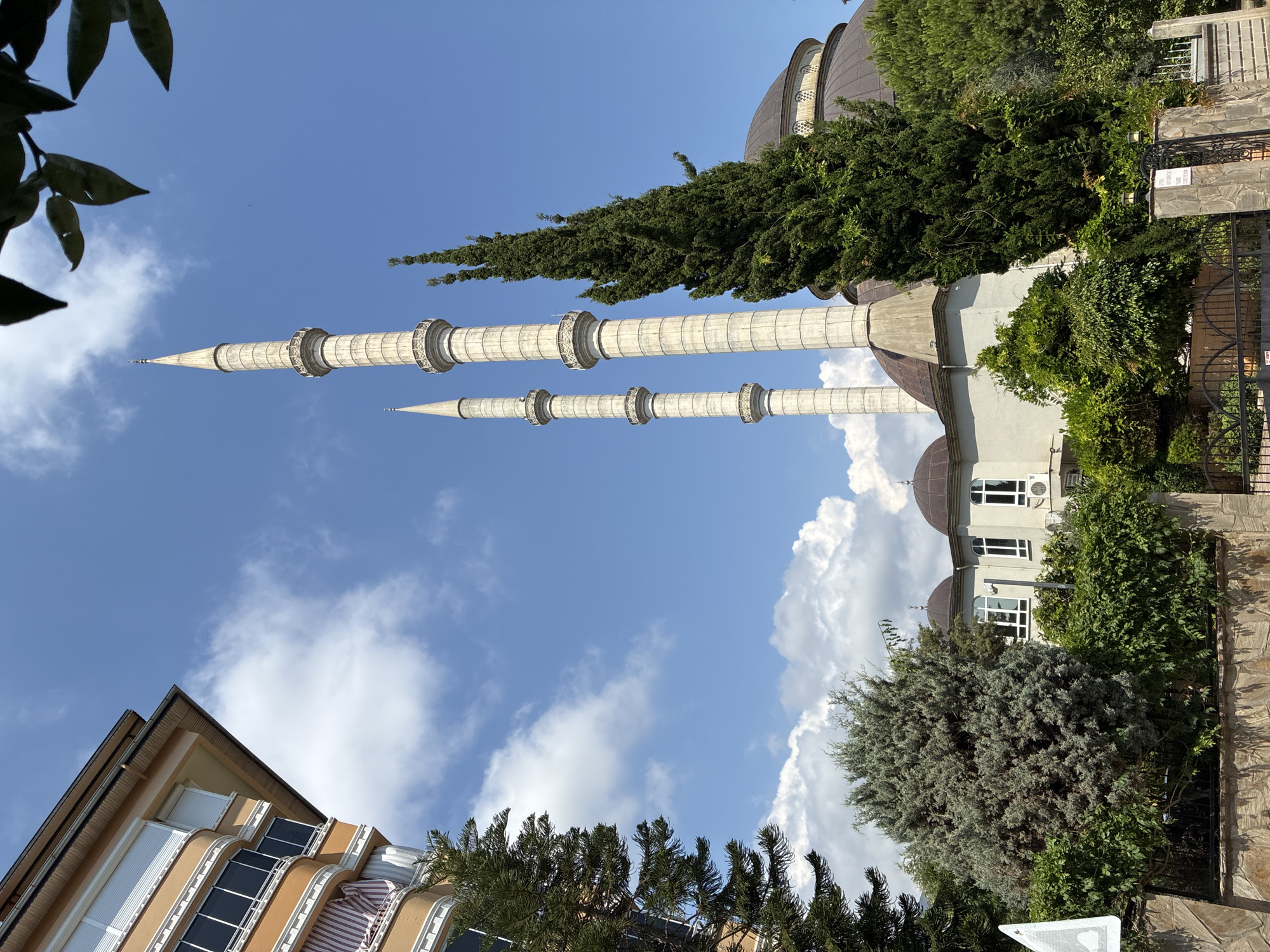 The image depicts two tall, slender minarets of a mosque, their white, ribbed structures reaching towards a bright blue sky dotted with white, fluffy clouds. The minarets have intricate decorative bands and pointed finials. Behind the minarets, the dome of the mosque and parts of its light-colored building are visible, partially obscured by lush green trees and foliage.
In the foreground and to the left, a section of an apartment building with a unique architectural style is visible. This building features a light brown facade with a curved design, multiple windows with dark frames, and what appear to be awnings or balconies.
The scene is set outdoors, likely during the daytime due to the bright sunlight casting shadows and illuminating the sky. The weather appears to be clear and pleasant, with a few clouds suggesting a warm day. The overall impression is a peaceful and serene view of religious architecture integrated with residential buildings and abundant greenery.
There is no visible text in the image. The location is identified as Alanya, Turkey.
