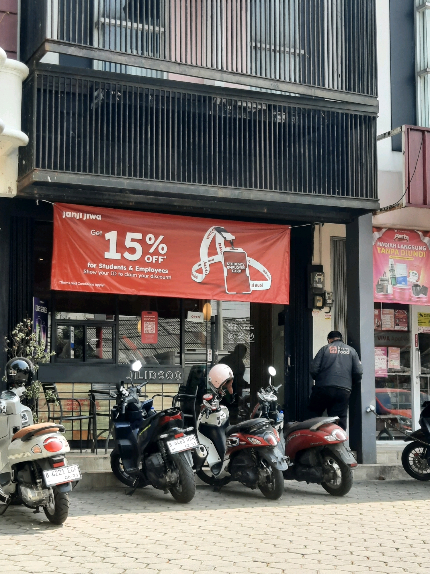 The image depicts the exterior of a coffee shop called "Janji Jiwa" located at Jilid 900 in East Jakarta, Indonesia. A large red banner hangs across the storefront, advertising a "15% OFF" discount for students and employees, with a requirement to show their ID. Several parked motorbikes are visible in front of the shop, suggesting a common mode of transportation in the area. A man wearing a dark jacket with a "Shopee Food" logo is standing near the entrance. The building appears to be a multi-story structure with dark metal railings. Adjacent to the coffee shop is a pink-themed storefront, possibly a pharmacy or convenience store, with promotional posters displayed. The ground is paved with hexagonal tiles, and some potted plants are placed near the entrance. The overall scene suggests a busy urban environment, likely during daylight hours given the lighting conditions.