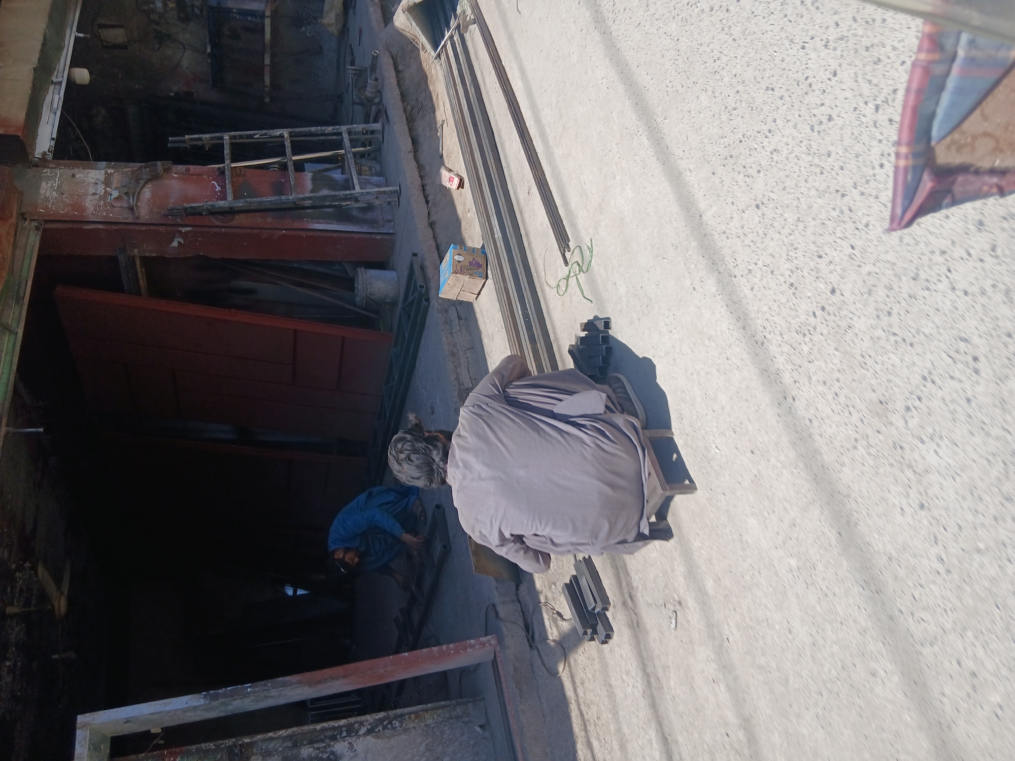 The image shows an outdoor scene in Quetta City Tehsil, Pakistan. Two men are present, one wearing a blue shirt and the other wearing a grey robe. The men appear to be working on or near metal bars. Objects visible in the scene include metal bars, a box, and several pieces of square metal tubing. The setting appears to be a roadside or area near a building. The ground is paved. The image is taken from a slightly elevated perspective.