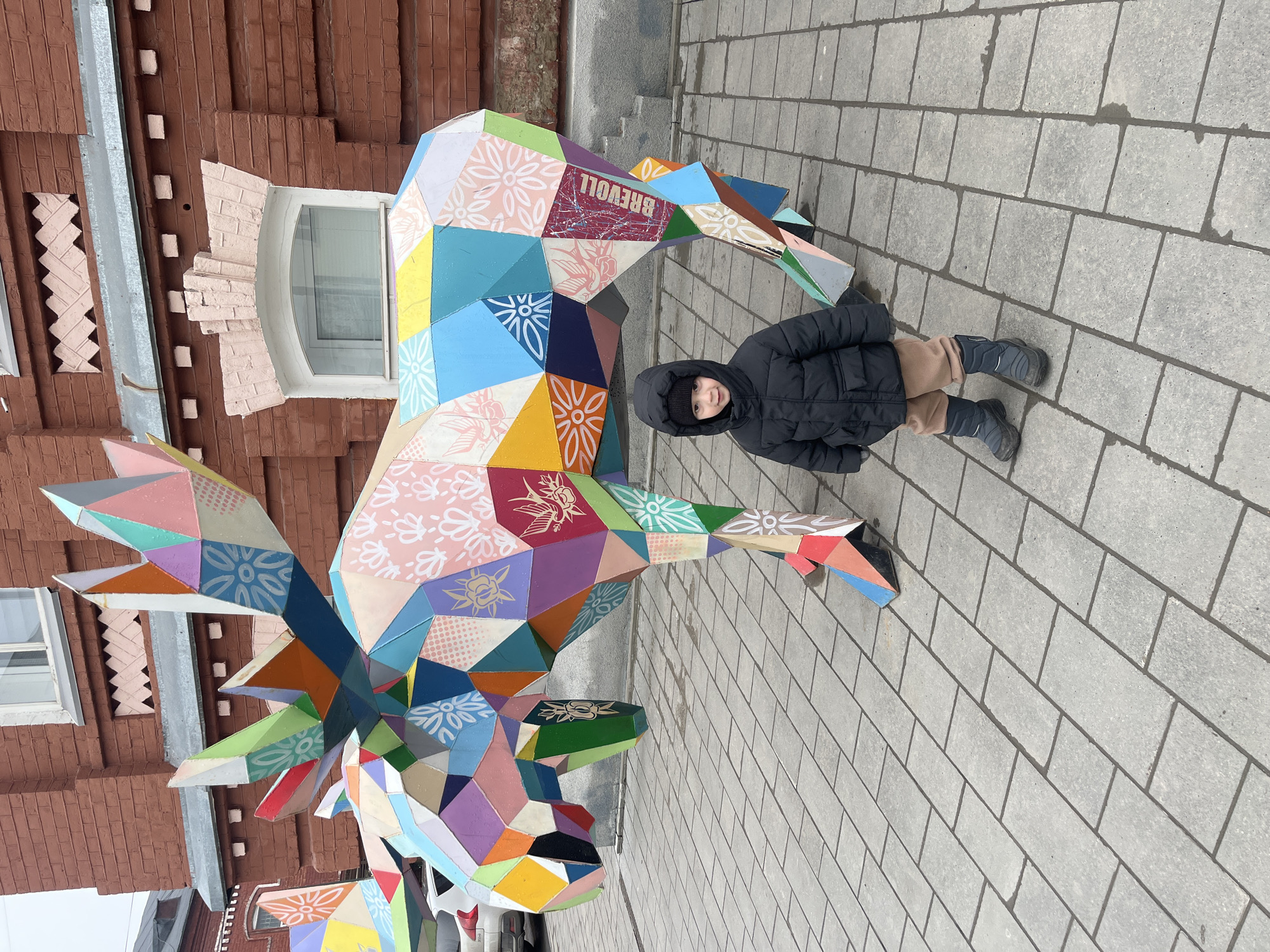 A young child wearing a black winter coat, tan pants, and black boots stands beside a colorful, polygonal animal sculpture. The sculpture, which resembles a moose or deer with large antlers, is constructed from numerous geometric shapes decorated with floral and other patterns. The sculpture has the text "BREVOLL" printed on it. The scene is set on a paved walkway made of light gray rectangular blocks. Behind the sculpture, a brick building with a window and decorative brickwork is visible. The photo was taken in the city of Ulyanovsk, Russia.