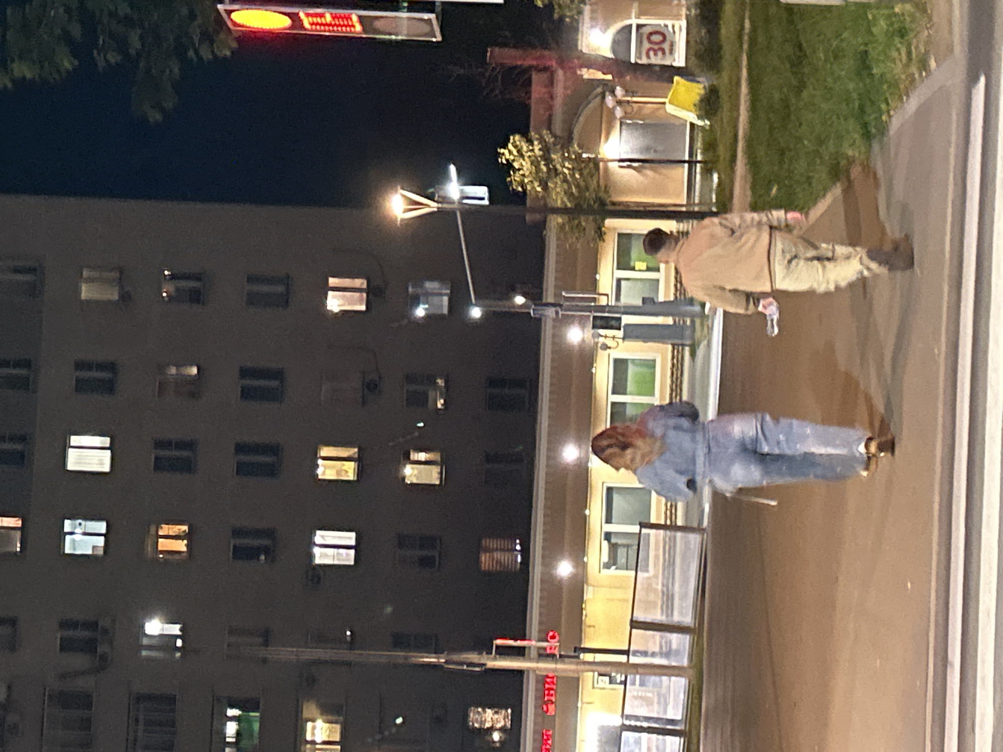 Two individuals, a woman and a man, are walking away from the camera on a paved pedestrian path adjacent to a grassy area. The woman is on the left and is wearing a denim jumpsuit. The man is on the right and is wearing a light brown jacket and camouflage pants. He is carrying a transparent bottle and a long object, possibly a stick or a broom.
In the background, there is a tall, multi-story building with numerous lit windows. Below the building is a lower structure with illuminated storefronts and windows. A street lamp is visible between the two people and the building. Traffic lights and signage are also present in the background, and a red and yellow sign with the number "30" is visible on the right side of the frame. The scene appears to be in an urban environment at night. The presence of Cyrillic text on the storefronts indicates the location is likely in Russia. The architectural style of the buildings and the visible signage are consistent with an urban area in Moscow, Russia.