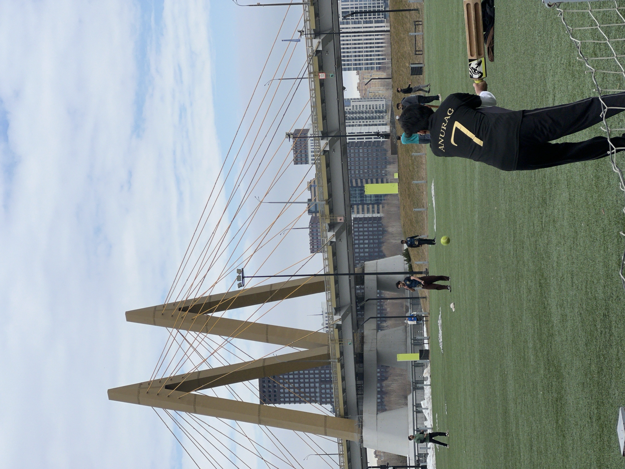 The image depicts a soccer field in Kazan, Russia, with the Millennium Bridge in the background. Several people are playing or watching the game. A person wearing a black shirt with the name "ANURAG" and the number "7" on the back is holding a cricket bat. A soccer ball is visible on the field. The Millennium Bridge is a prominent architectural structure in the background, with its distinctive cable-stayed design. Buildings and a cloudy sky are also visible.