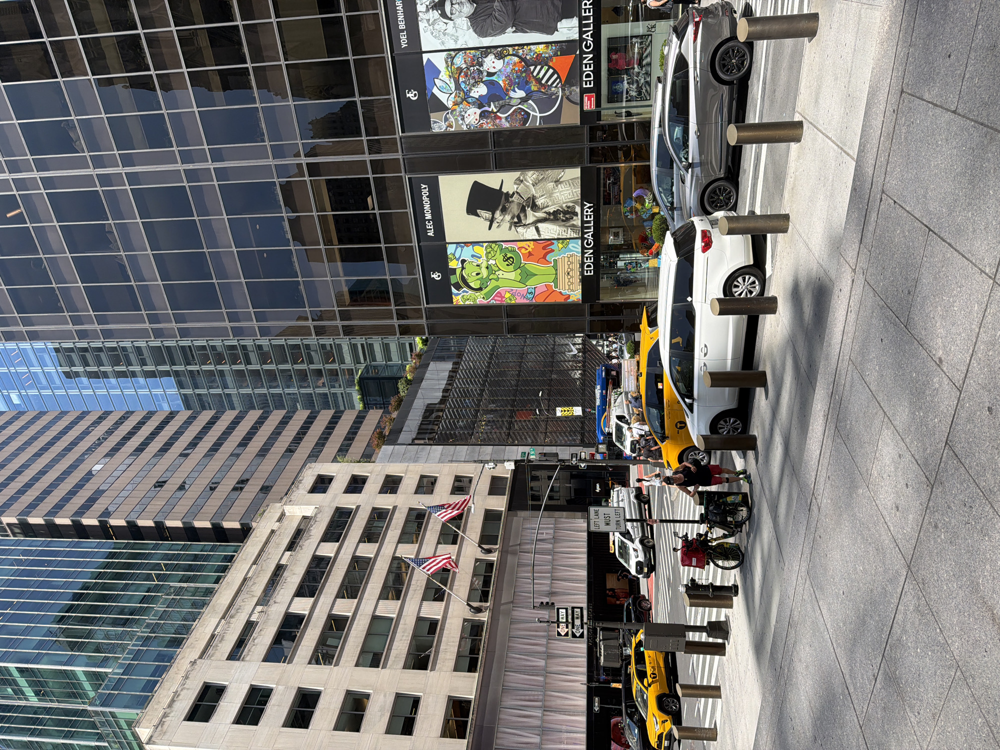 This is a medium-angle, outdoor, vertical shot of a bustling street scene in New York City. The sun is shining, casting sharp shadows, suggesting it's a bright, clear day, likely midday. The focus is on the street and sidewalk in the foreground, with tall buildings dominating the background.
Several vehicles are present, including a white minivan, a gray SUV, and multiple yellow taxis, indicating a busy urban environment. Some of these vehicles are parked, while others appear to be in motion or waiting. People are visible on the sidewalk, some walking, others perhaps waiting for a taxi or observing the scene. There are also bicycles parked along the sidewalk.
The buildings showcase distinct architectural styles. One building on the left has a modern glass facade, reflecting the sky and surrounding structures. Another building, more centrally located, features a lighter-colored stone exterior with numerous windows and American flags flying from poles. Further back, another building displays artwork on its exterior, with prominent signage for "EDEN GALLERY" and names like "ALEC MONOPOLY" and "YOEL BENHAR". This suggests the area is a commercial or artistic district.
Visible text includes street signs like "ONE WAY" and "LEFT LANE MUST TURN LEFT," as well as gallery names. The overall impression is a lively, everyday moment in a dense urban landscape.