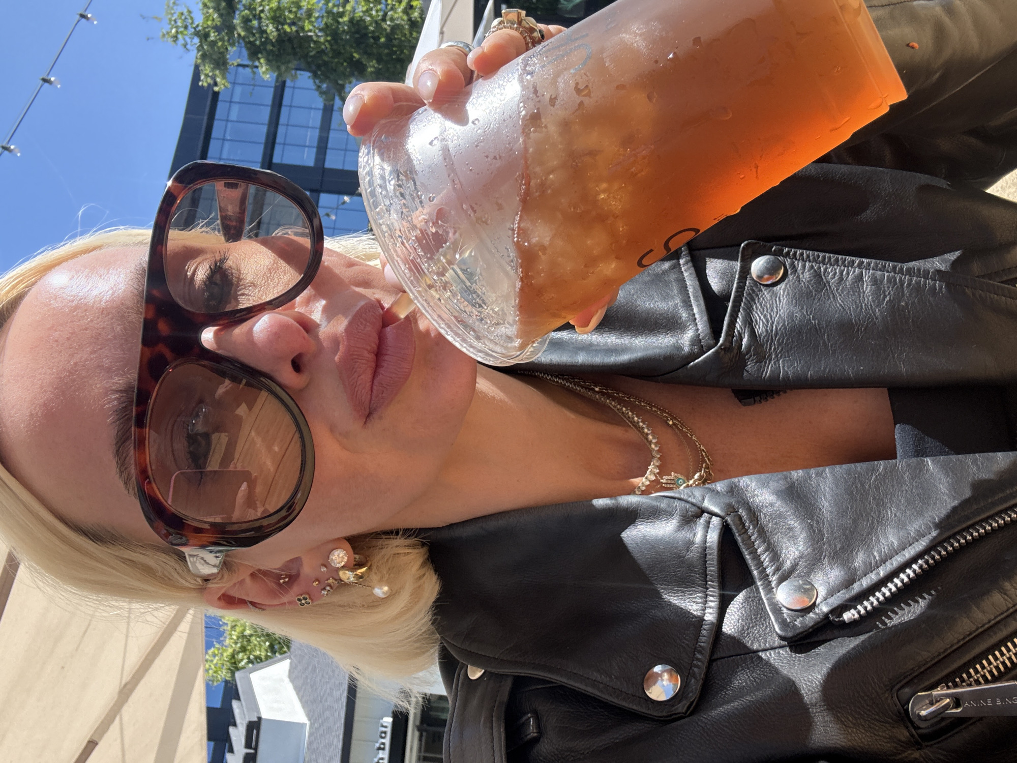 A person, likely female, with blonde hair, is captured from a low, upward-looking angle, appearing to sip from a large plastic cup containing an amber-colored iced beverage. Her lips are visible, adorned with pink lipstick, and her face is partially obscured by large, tortoiseshell-patterned sunglasses.
She wears a black leather jacket with silver snap buttons and zippers, one of which prominently displays the brand name "ANINE BING". Around her neck, several gold-toned necklaces are visible, including one with a small charm, possibly an evil eye or Hamsa hand. Her left ear is accessorized with an array of earrings, including a large diamond stud, a pearl drop earring, and several smaller decorative studs. Multiple rings are worn on the fingers of her right hand, which holds the drink. The plastic cup shows condensation and has a partially visible, stylized 'S' or similar letter as part of a logo.
The scene is set outdoors on a bright, sunny day, indicated by clear blue skies and strong lighting. In the background, lush green tree foliage and a modern building with dark glass windows are visible, reflecting the sunlight. String lights stretch across the upper portion of the frame. To the far left, a light-colored awning or umbrella structure is partially seen, and a distant sign shows the text "h bar". The overall atmosphere is casual and relaxed, suggesting a moment of enjoyment in an urban outdoor setting in San Diego, USA.