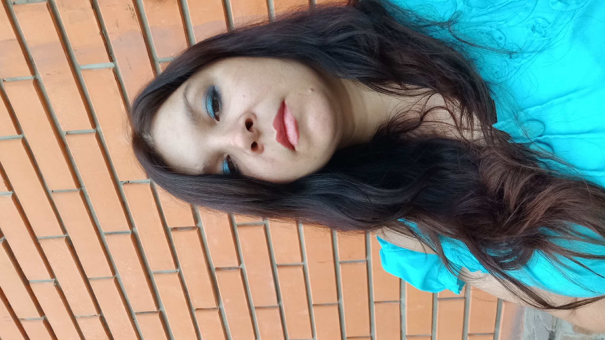 A woman is photographed from the chest up, with her face tilted slightly downward and to the left. She has dark brown hair that falls around her shoulders and a bright turquoise top. Her makeup includes blue eyeshadow and light pink lipstick. She is standing in front of an orange brick wall. The image is likely taken outdoors, possibly in a public space within the city of Kropyvnytskyi, Ukraine.