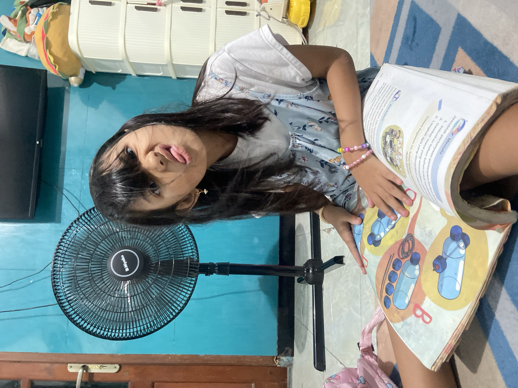 A young girl with dark hair sits in a room, sticking her tongue out at the camera. She is wearing a light blue dress with a white t-shirt underneath and a colorful bracelet on her wrist. She is holding a large book open in front of her, displaying colorful illustrations. Behind her is a standing fan and a blue wall with a wooden door frame near the bottom. The room appears to be in a home, and the text in the book is written in Indonesian. This is in the city of Gelam, Indonesia.