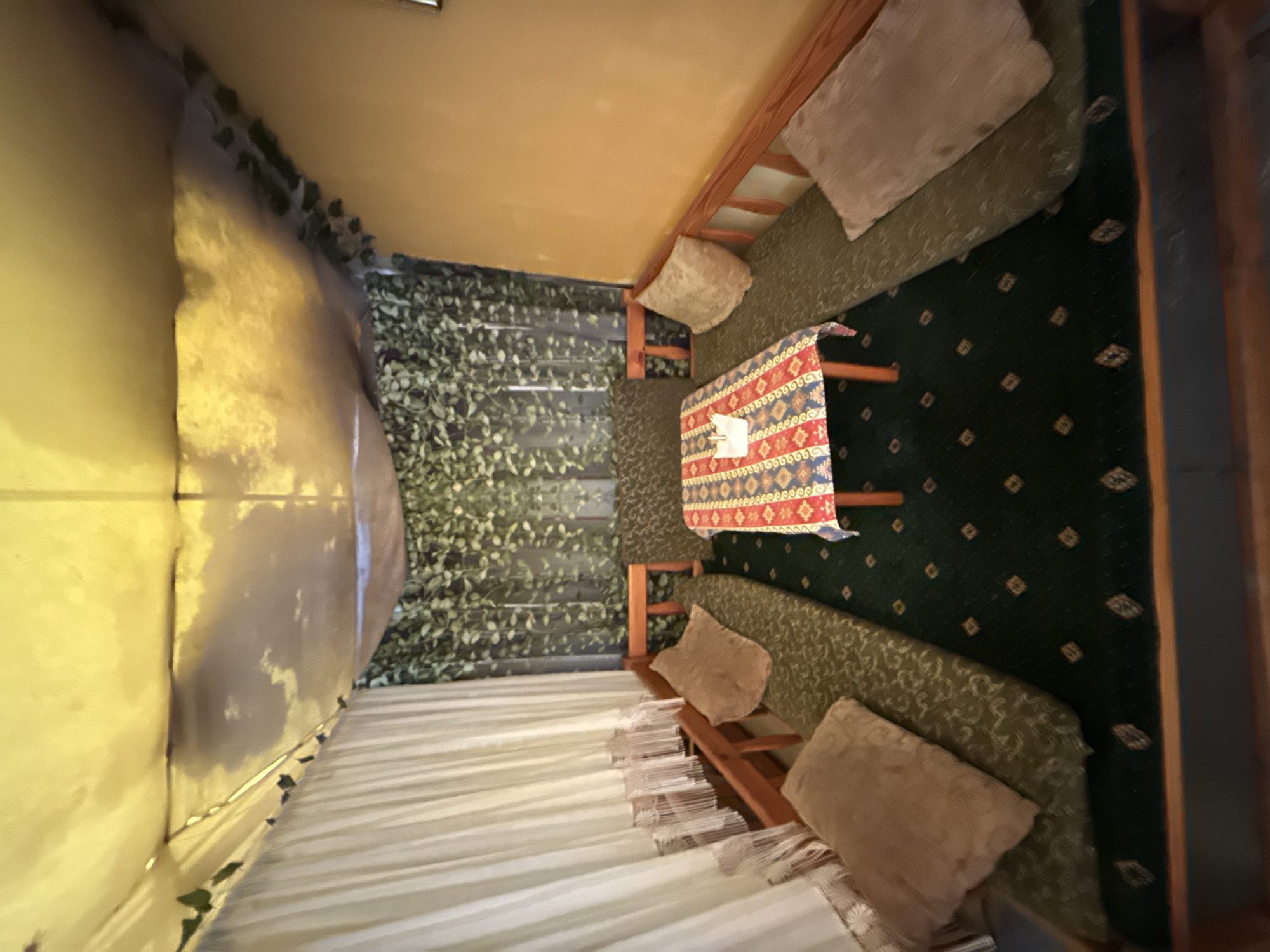 A high-angle shot depicts an interior space. The room is divided into distinct sections. On the left, a portion of a tent-like structure with light-colored fabric walls is visible. Next to it, artificial vines cover a wall, providing a green, leafy backdrop. Towards the center of the image, there's a table covered with a patterned cloth. Seating areas are positioned on opposite sides of the table. One seating area features cushions on a bench, while the other side has a more structured bench with cushions and pillows. The floor is covered with a dark green rug adorned with a repeating pattern. The room seems to blend elements of an outdoor and indoor setting. The exact location within the city of Unknown, Unknown is not ascertainable from the visual information provided.