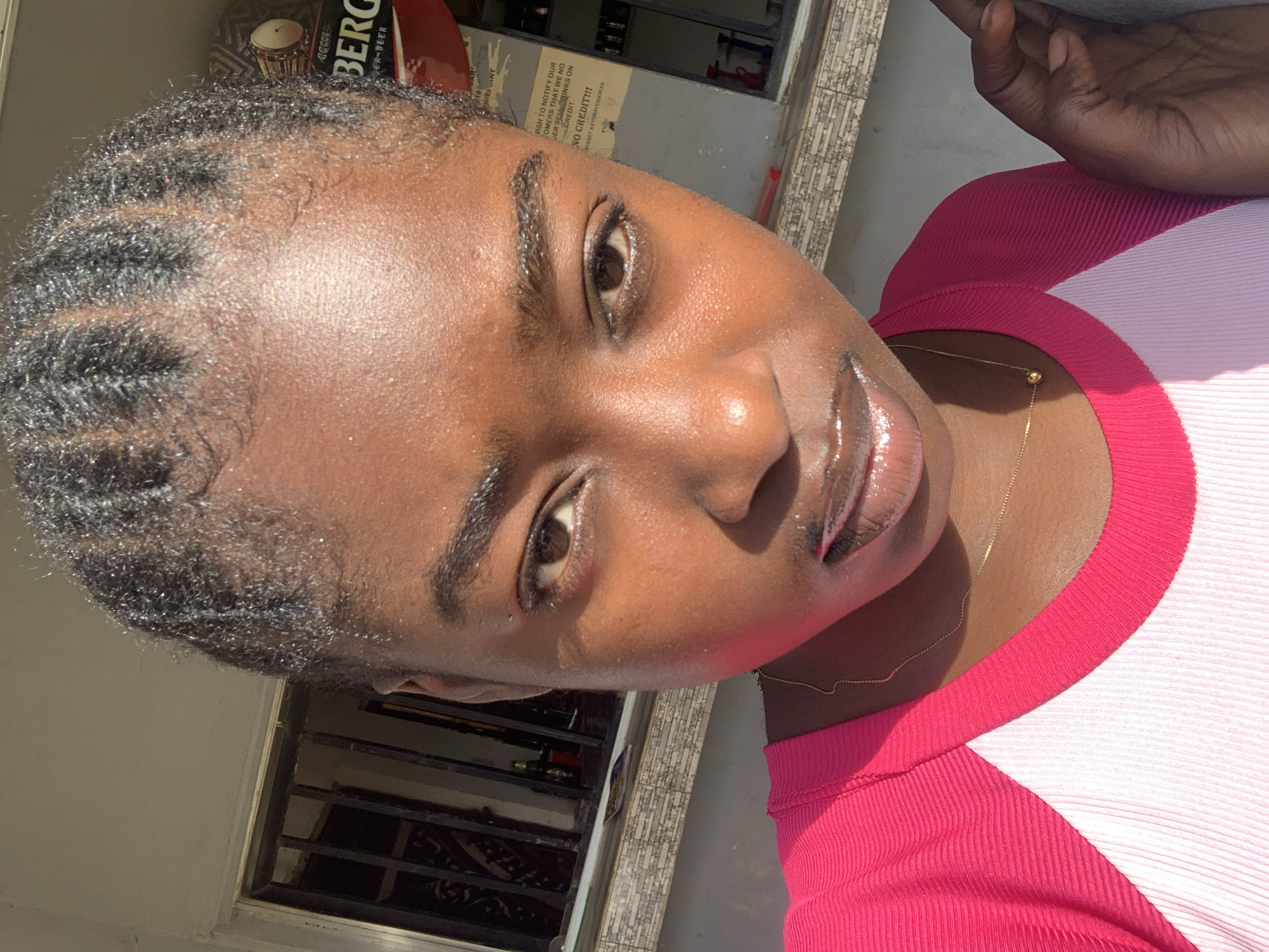A close-up shot depicts a dark-skinned woman with braided hair wearing a pink and white top. She has full lips with a glossy finish and is wearing a delicate gold necklace. The background shows a window, an advertisement with the word "BERG" visible, and part of a wall or exterior setting. The location is New Stadium, Nigeria.