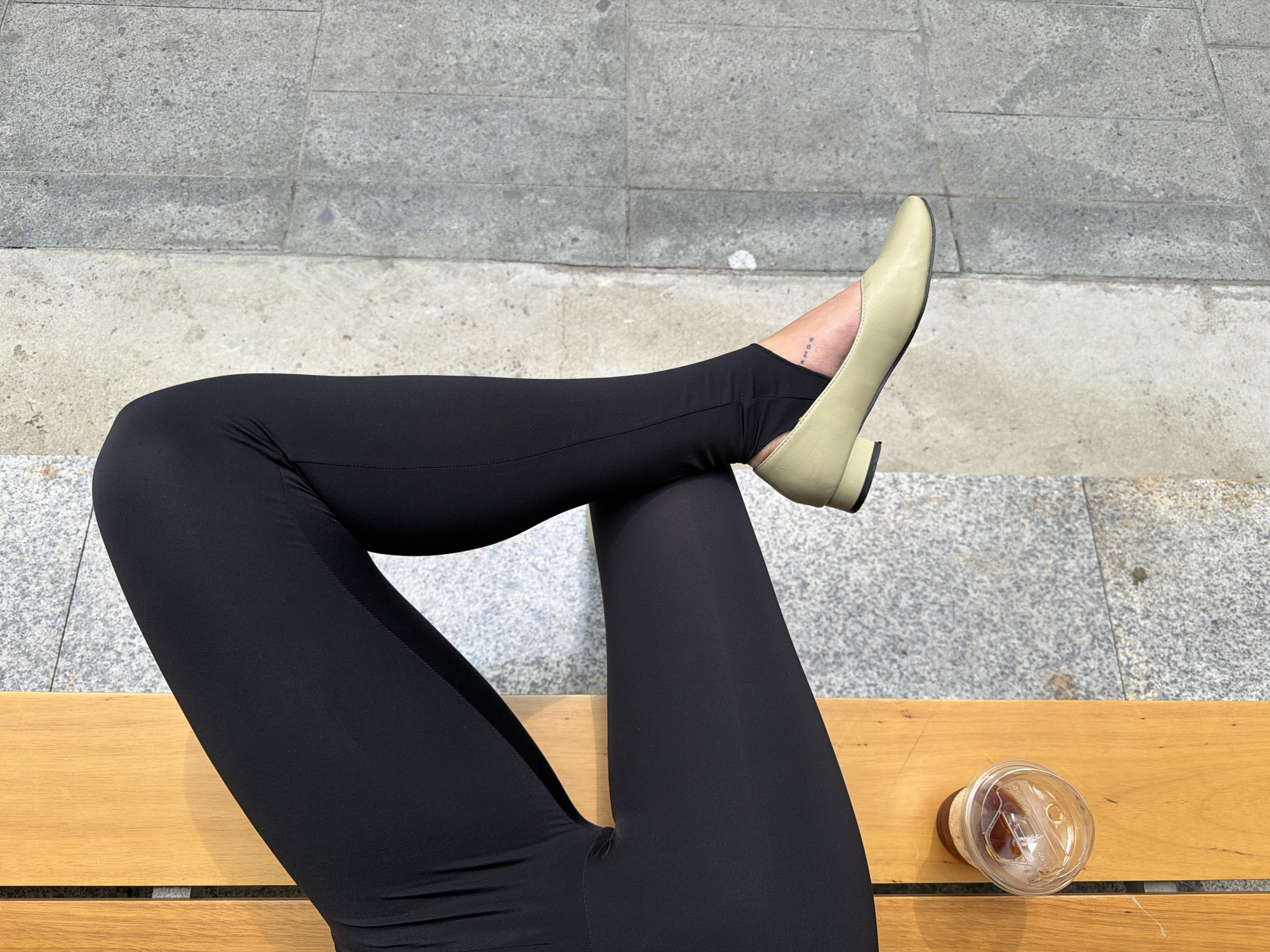 A woman wearing black leggings and light green shoes sits on a wooden bench in Seoul, South Korea. Her legs are crossed at the knees. A disposable plastic cup containing a beverage is on the bench beside her legs. The ground beside the bench is made of gray concrete tiles. The woman has a small tattoo on her ankle.