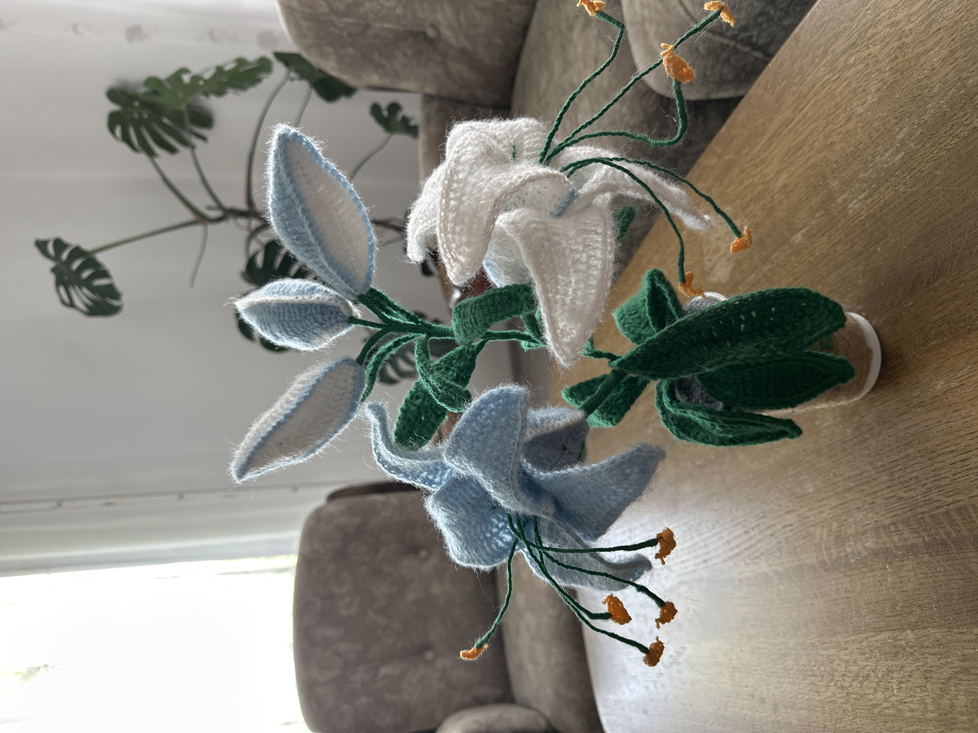 An indoor still life features a crochet arrangement of white and light blue lilies with green leaves and orange stamen, placed in a small pot on a wooden table. In the background, two upholstered chairs and a large-leafed houseplant are visible. The scene is illuminated by natural light filtering through a window on the left.