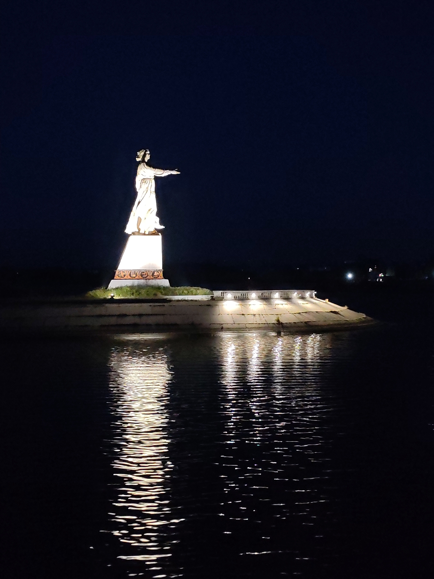 The photograph captures a nighttime scene featuring a illuminated statue of a woman standing on a raised platform beside a body of water. The statue, which appears to be made of stone and is lit from below, is depicted with its right arm extended. The platform is also illuminated with lights along its edge, and these lights cast reflections on the dark water. In the background, distant lights suggest the presence of buildings or other structures. This monument is the "Motherland" statue, located on the embankment of the Volga River in Rybinsk, Russia.