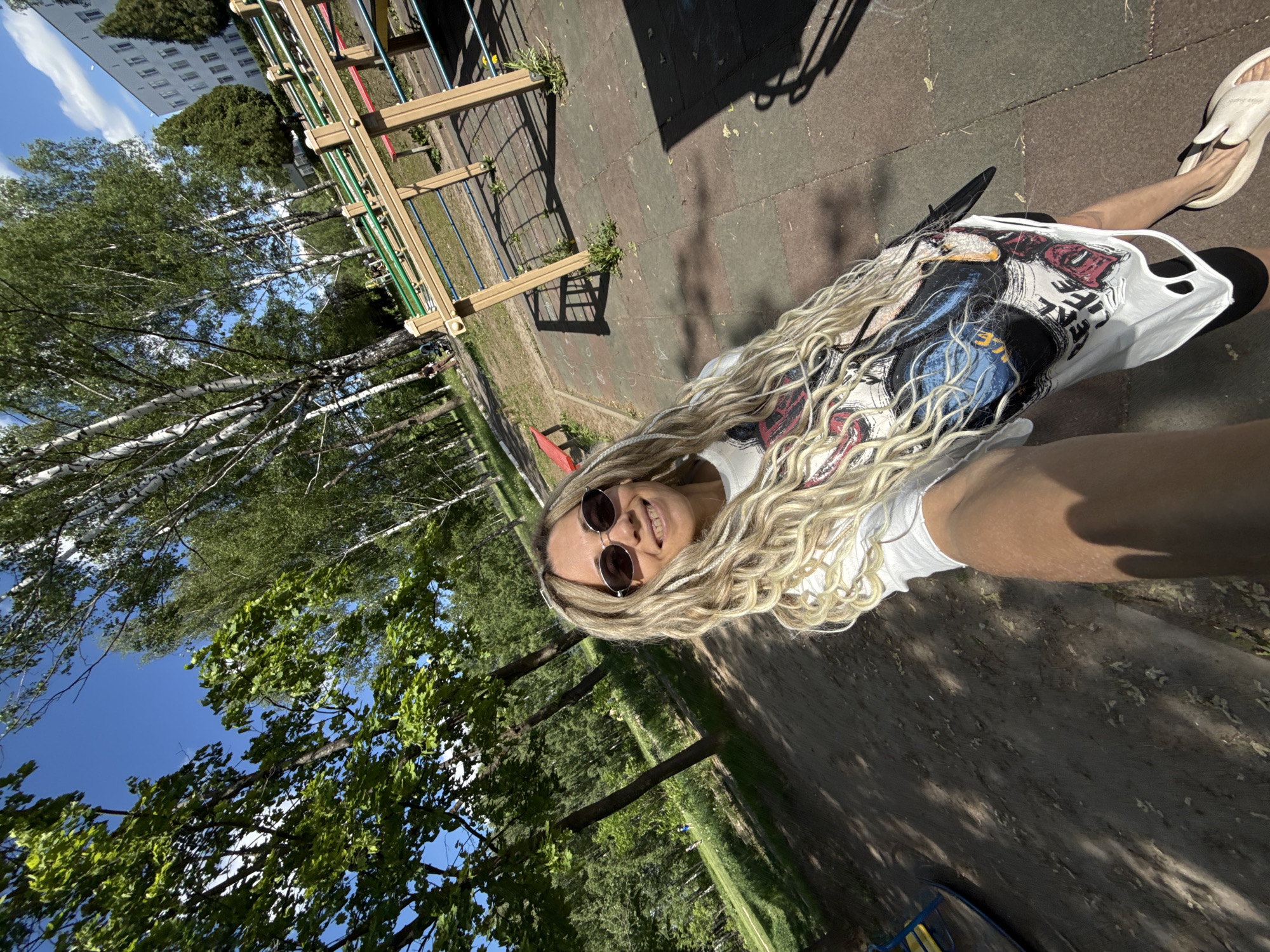 The image shows a woman with blonde braids wearing sunglasses and a white t-shirt with a graphic print. She is outdoors, likely in a park or playground, based on the presence of play equipment in the background. The ground is paved and there are trees visible in the background. The location may be Шебунино, Russia, as requested.