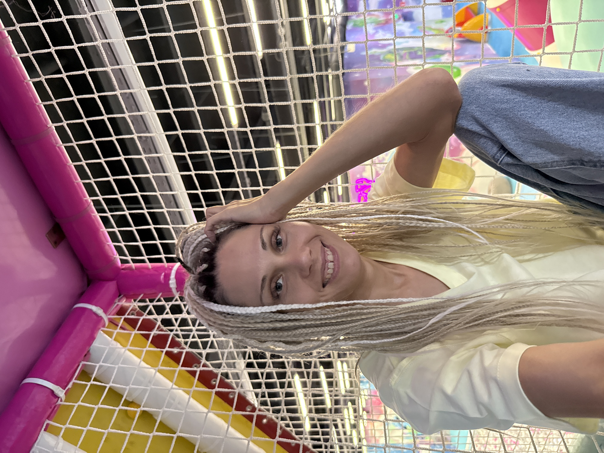 A medium shot captures a woman with blonde braided hair, wearing a light yellow shirt and blue jeans, sitting inside a colorful indoor playground. The background is a netting structure with bright pink and yellow poles, and further beyond, soft play equipment is visible. The woman is smiling and has her hand resting on her forehead. The image does not provide any information about the city of Zhukovo, Russia.