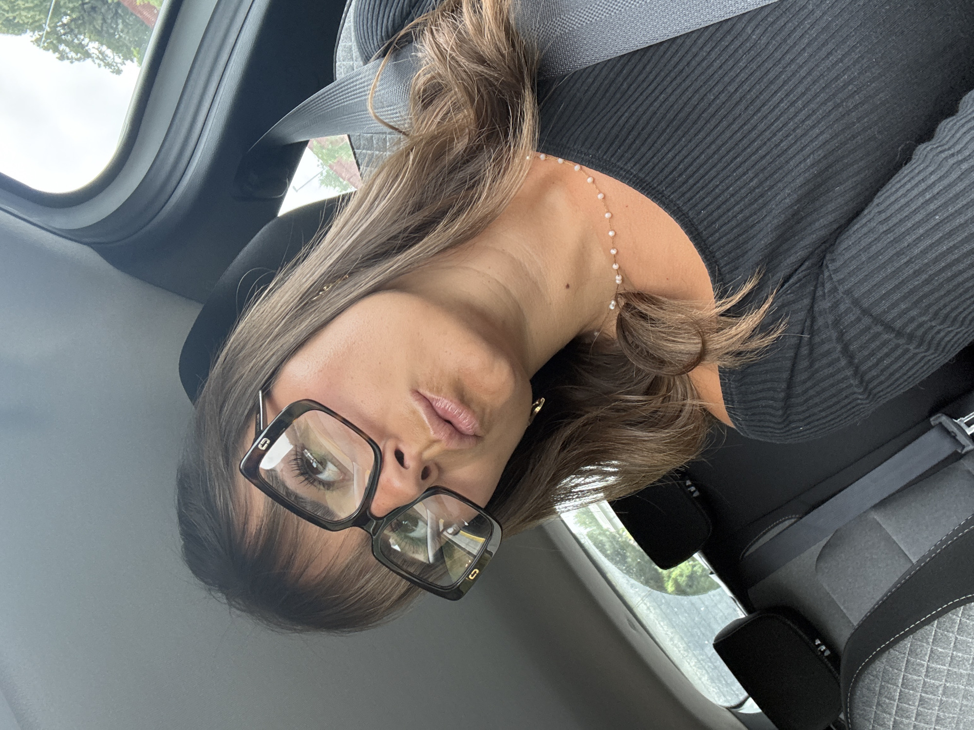 A woman with long brown hair and wearing black-rimmed glasses is in a car. She is wearing a black ribbed top and a necklace with small pearls. She has a seatbelt on. The car's interior is visible, with a gray seat and a window showing trees outside. The location is not discernible as being Saint-Laurent-du-Var, France based solely on this image.