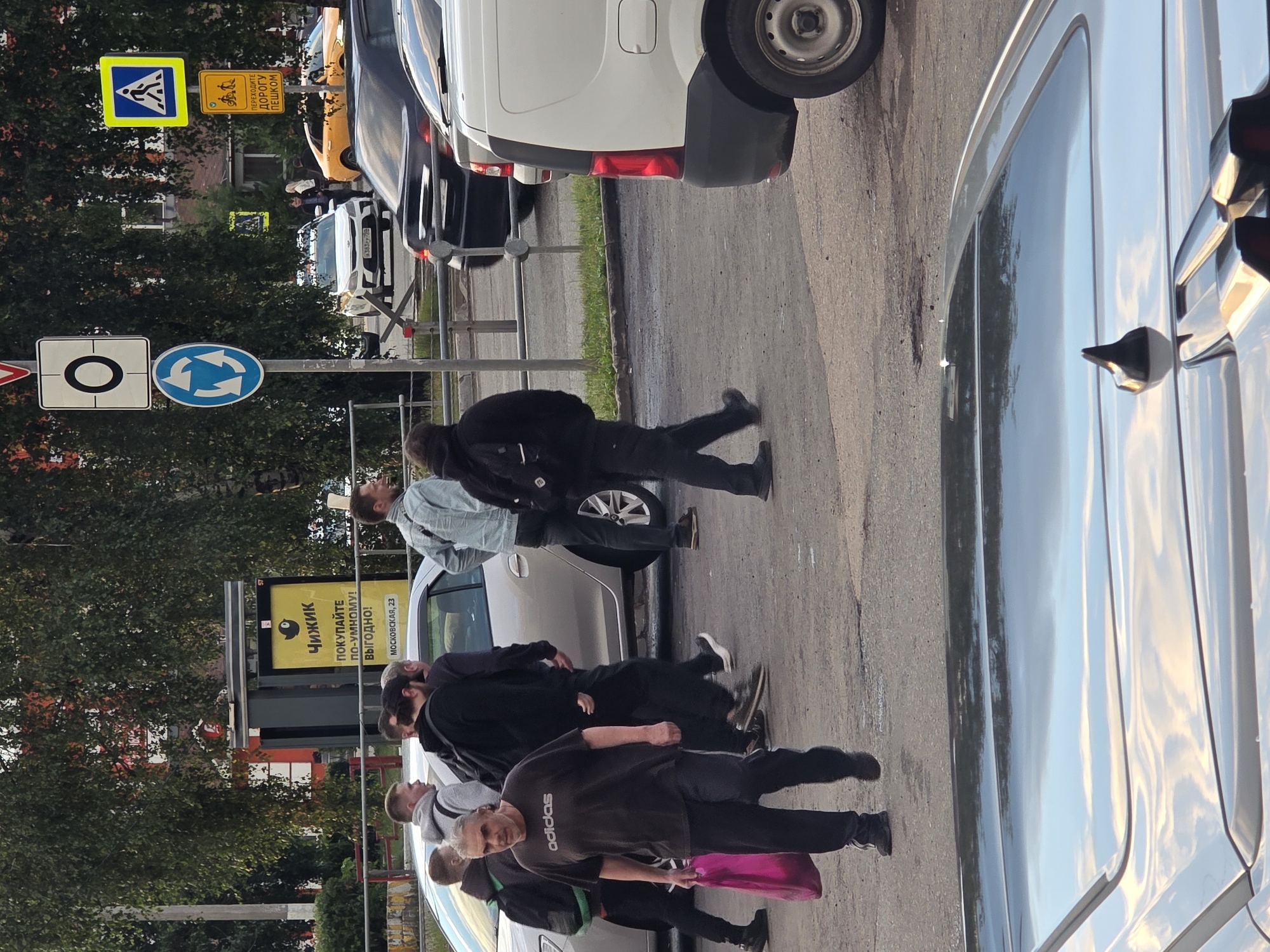 In an outdoor urban setting, several individuals are walking near a street with traffic. A man in a dark t-shirt and dark pants, carrying a pink bag, walks towards the camera. Behind him, a group of men, some wearing hoodies and backpacks, are also moving. A silver car is parked on the left, and other vehicles, including a white van and a yellow car, are visible further down the street. Traffic signs, including a pedestrian crossing sign, a roundabout sign, and a sign with a circle and a dot, are present. The ground is paved, and there are trees and a building in the background. The lighting suggests it is daytime, possibly late afternoon.
