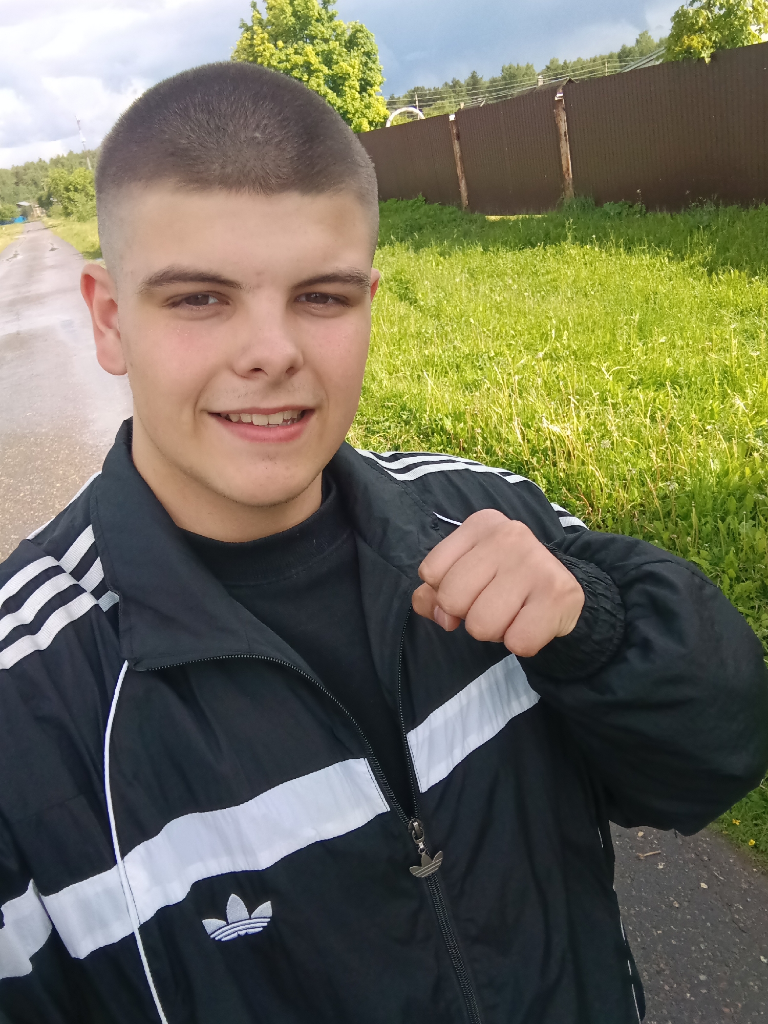 A young man stands on a paved road in Красный Маяк, Russia. He is wearing a black Adidas jacket with white stripes and an Adidas logo. He has short, cropped hair and is smiling. In the background, there is a green field, a wooden fence, trees, and a cloudy sky. The road appears wet.
