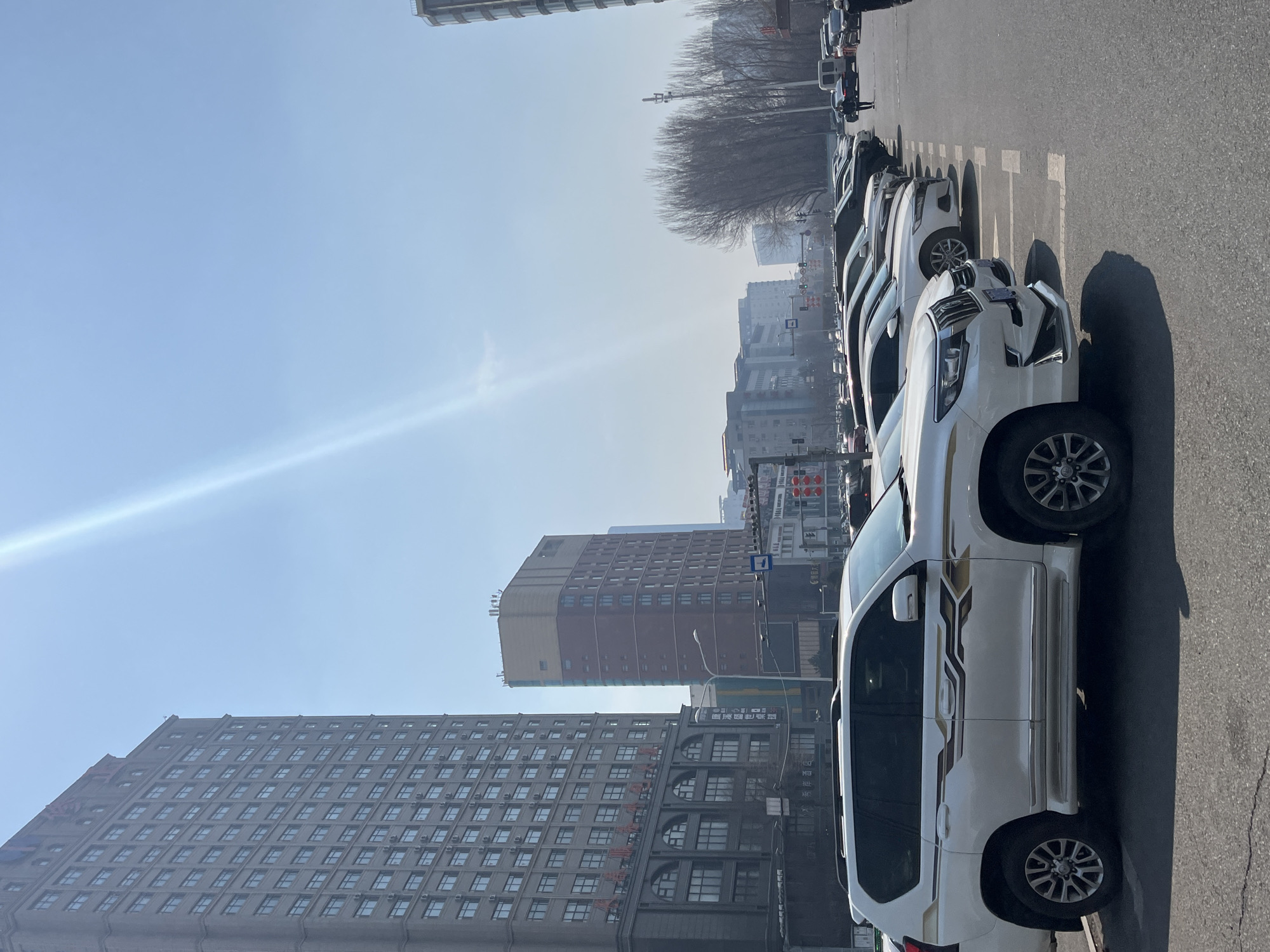 A daytime outdoor shot shows a row of parked white SUVs lining a street in Changchun City, China. The vehicles fill the right side of the frame and cast strong shadows on the asphalt. Behind the vehicles, a large grey brick building with numerous windows extends to the left, alongside a brown and tan building. Bare trees are visible in the distance. The sky is blue with a faint haze. A few other vehicles are also visible further down the street.