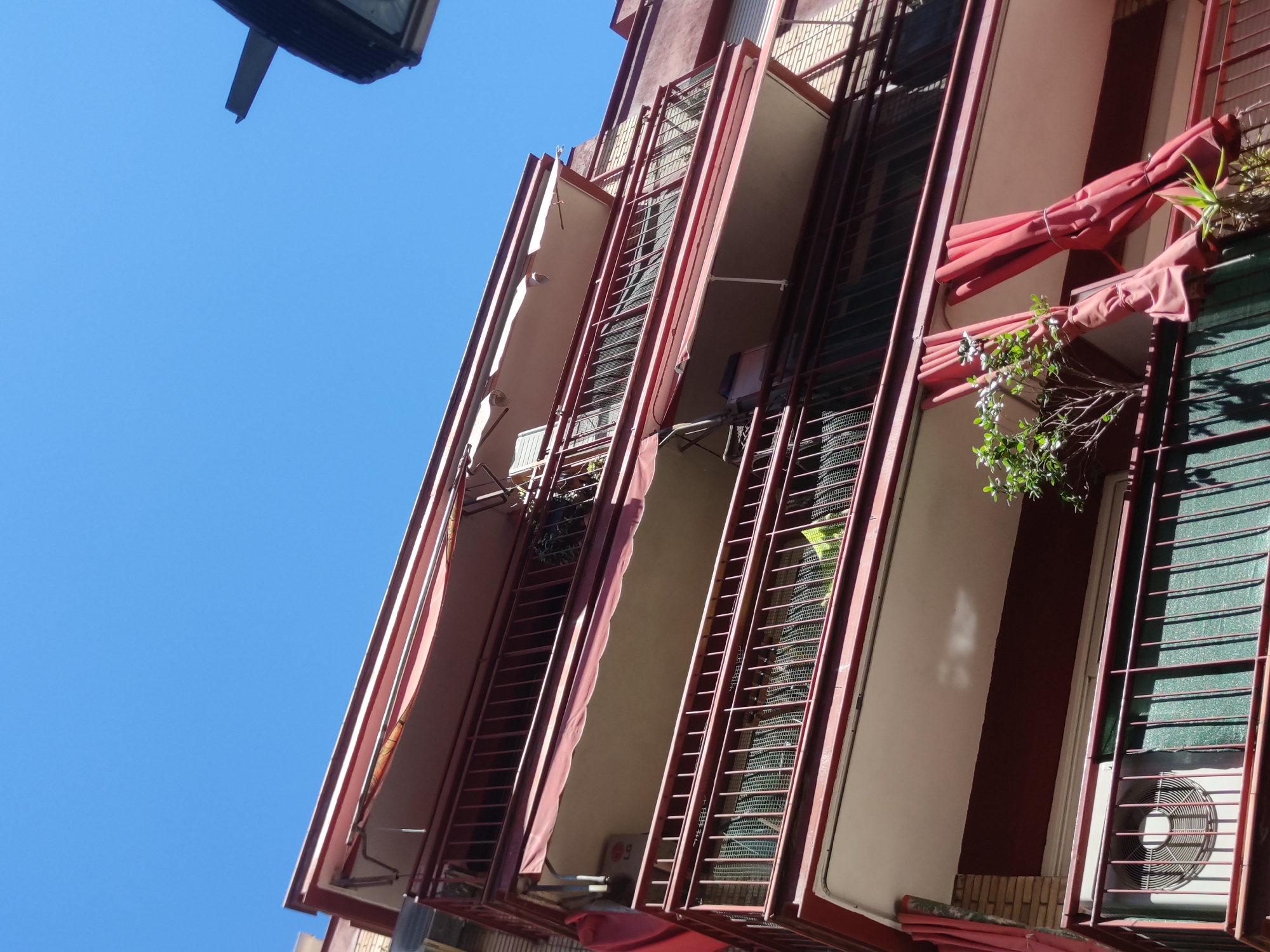 The image shows the exterior of a multi-story building in Unknown. The building features a red and white color scheme and has enclosed balconies with metal bars. Some balconies have rolled-up awnings and plants. An air conditioning unit is visible on one of the balconies. The sky is clear blue, and there is a street light in the upper corner of the image.
