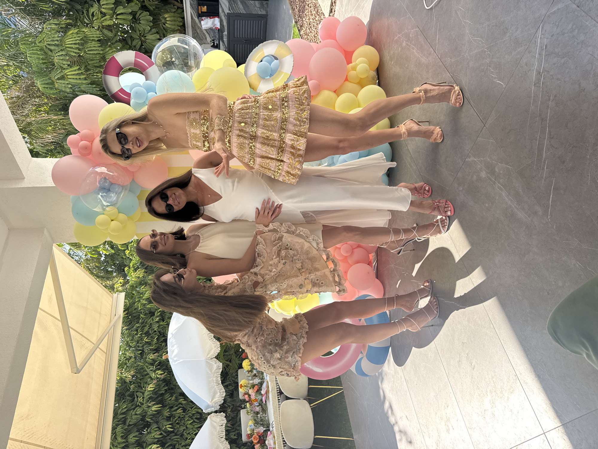 Four women are posing for a photo outdoors, in a paved courtyard area with greenery in the background. A table with floral arrangements is visible to the left, and a collection of colorful balloons is behind the women. The woman on the far right is wearing a strapless pink dress with gold embellishments and sunglasses. The woman next to her, second from the right, is wearing a long white dress and has her hand on her pregnant belly. The woman second from the left is wearing a light-colored dress with intricate details and sunglasses. The woman on the far left is also wearing a light-colored dress with similar details and sunglasses. The overall scene appears to be a celebration or party. The location is Agios Tychona Community, Cyprus.
