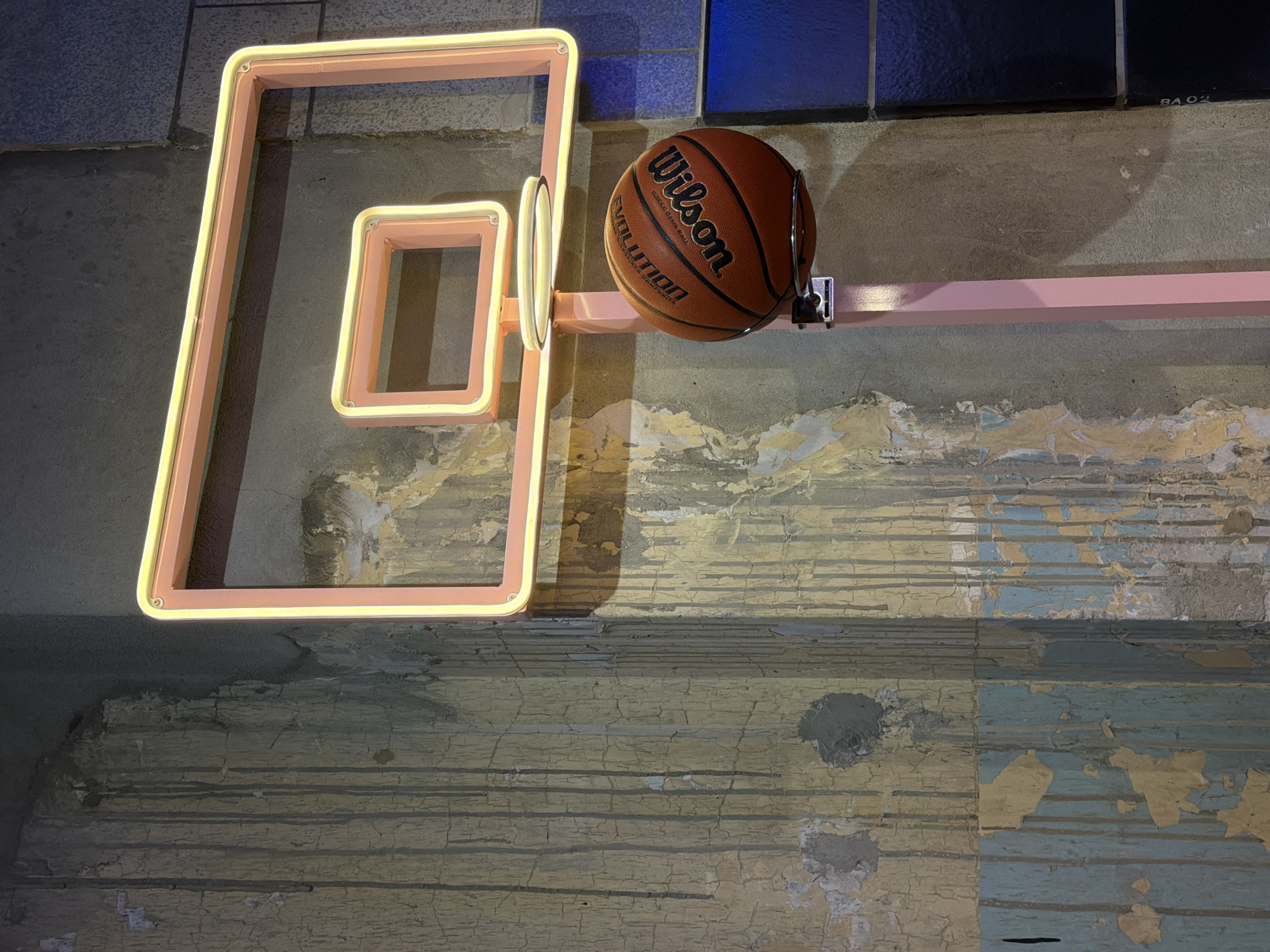 A basketball rests on a pink horizontal bar mounted on a wall, against a backdrop of peeling paint and geometric neon light fixtures. The basketball is an orange-brown color and the name "Wilson" is clearly visible on its surface. The pink bar extends from the basketball towards the right edge of the frame. Above the bar, there are tiles of navy, black, and grey. The wall itself is comprised of horizontal layers with sections of exposed and peeling light-yellow and blue paint. The geometric shapes on the left of the image are pink frames that contain neon lights, which appear to be a decorative element of the scene.
The lighting is provided by the neon fixtures and possibly an external light source. The neon light illuminates the wall above the bar and causes shadows on the peeling paint. The exact location in London, UK, cannot be determined from the image. There is no indication of any people, actions, or interactions within the scene.