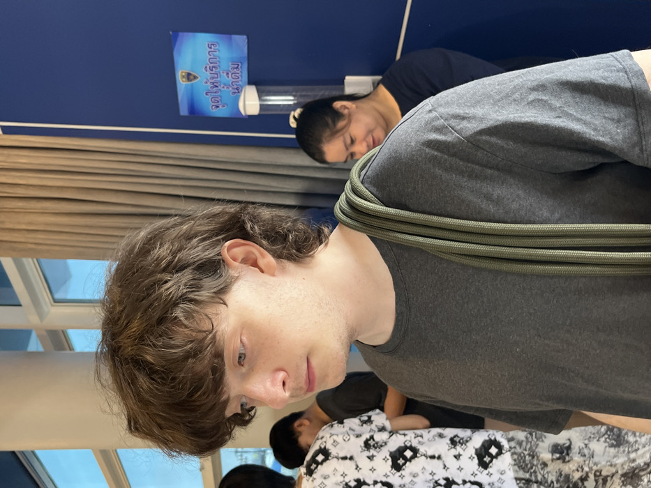 The image captures an indoor scene, appearing to be a casual gathering or waiting area, presented at a 90-degree counter-clockwise rotation from its natural upright orientation.
In the foreground, slightly to the right, a young male with light skin and shaggy, dark brown hair (styled somewhat like a mullet) is visible from the chest up. He is wearing a plain grey t-shirt. A thick, olive-green rope is coiled several times and draped over his right shoulder and neck. He is looking downwards and slightly to his left, with a contemplative or subdued expression.
Behind and to the left of the foreground male, another person, an adult female, is partially visible. She has darker skin and dark hair pulled back into a ponytail. She appears to be resting or asleep, with her head tilted downwards, and is wearing a dark-colored top.
Further in the background, several other blurry figures are present, suggesting more people are in the vicinity. One figure, lower right, is wearing a white shirt with repetitive black patterns, possibly a logo or design, though it's too indistinct to confirm.
The background features a solid blue wall. Mounted on this wall, above the female figure, is a transparent dispenser, likely for water or hand sanitizer. Above the dispenser, a small blue sign is affixed to the wall. The sign features a golden emblem or crest at the top and Thai text below it. The text reads "จุดให้บริการ น้ำดื่ม", which translates to "Drinking water service point" or "Drinking water dispenser".
To the far left of the image, window blinds are visible, suggesting a window or glass partition is present, letting in some ambient light from outdoors, indicating it is likely daytime. The overall lighting is soft and indoor-typical. The atmosphere seems relaxed, with some individuals resting or waiting.