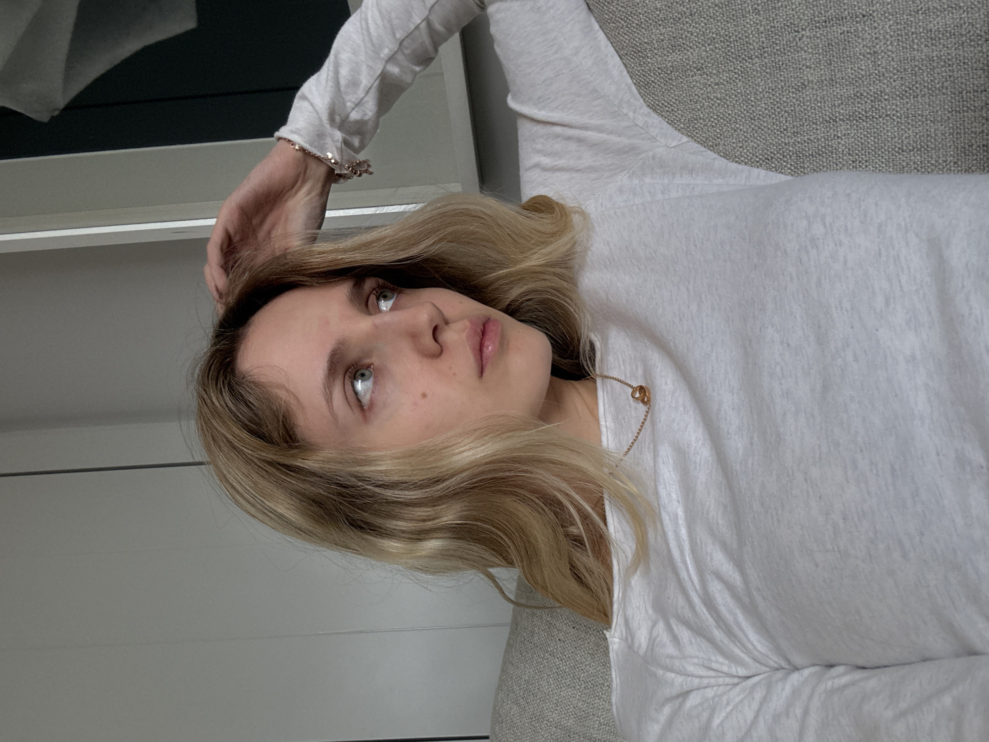 A woman with blond hair and blue eyes is lying down, wearing a white long-sleeved shirt and a gold necklace. Her left hand is raised to her head. She is on a gray surface, possibly a sofa, indoors. A white wall and a window or door frame are visible in the background. There is no indication of the location being in Mexico City.