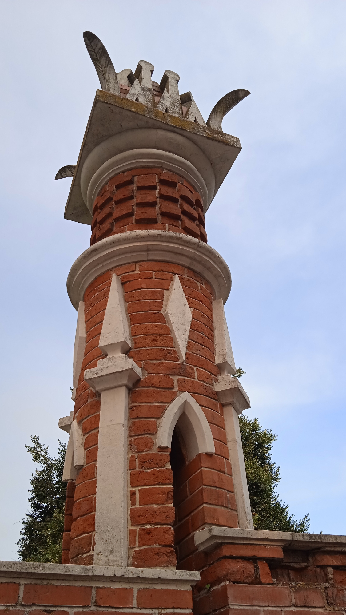 A low-angle, close-up shot focuses on the top section of a decorative brick tower against a pale blue sky. The tower is constructed from weathered red bricks and features lighter stone accents. The stone detailing includes a circular band below the topmost section, diamond-shaped insets, pointed vertical elements, and arched openings near the base. The very top of the tower has a square stone cap with a decorative, crenellated edge and stylized, bird-like or feather-like stone elements. Green foliage from trees is visible behind and to the sides of the tower. The image does not contain any people or indicate a specific location in Moscow.