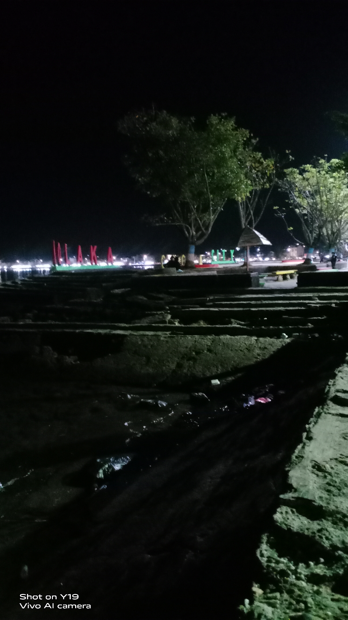 At night, the scene depicts the waterfront area in the city of Pamekasan, Madura, Indonesia. Large, illuminated red letters spelling "PAMEKASAN" stand out against a dark background. The letters are positioned along a walkway or embankment. In the background, there are trees, lights from buildings or streetlights, and several people are visible. One group of people is gathered near a small, thatched-roof shelter. The foreground shows tiered concrete structures, possibly steps or seating, leading down to a body of water. The water's surface is dark, reflecting some of the lights from the shore, and debris is visible in the water and along the edges of the structures. The overall impression is of a public recreational space illuminated for evening use.