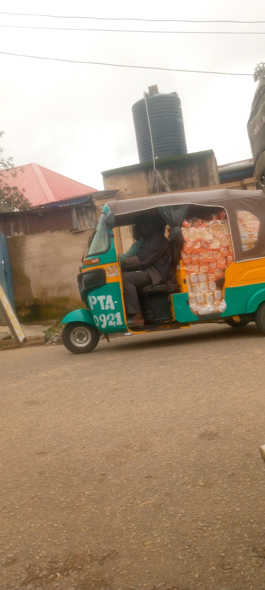 A three-wheeled auto rickshaw, painted green and yellow, is loaded with many loaves of bread. The auto rickshaw is being driven by a person wearing a dark jacket and dark pants. The vehicle has "PTA-0921" written on its side in white lettering. It is parked on a paved road. In the background, there is a building with a red roof, a water tank on top of a structure, and a stone wall. The sky is overcast.