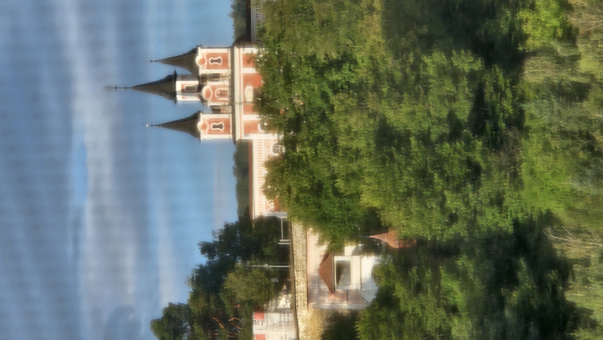 The image shows a view of the Church of the Holy Spirit in Prešov, Slovakia. The church is a prominent building with a red and white facade and three spires. It is situated on a hill surrounded by trees. The sky above is cloudy and blue. The photograph is taken from a distance, and the image is slightly blurred, giving it a soft, painterly quality. In the foreground, there is a dense canopy of green trees, obscuring the lower portion of the scene. The church's architecture is distinctly baroque, characterized by its ornate design and colorful exterior. The overall impression is one of historical significance and natural beauty, with the church serving as a focal point against the backdrop of lush vegetation and a dramatic sky.