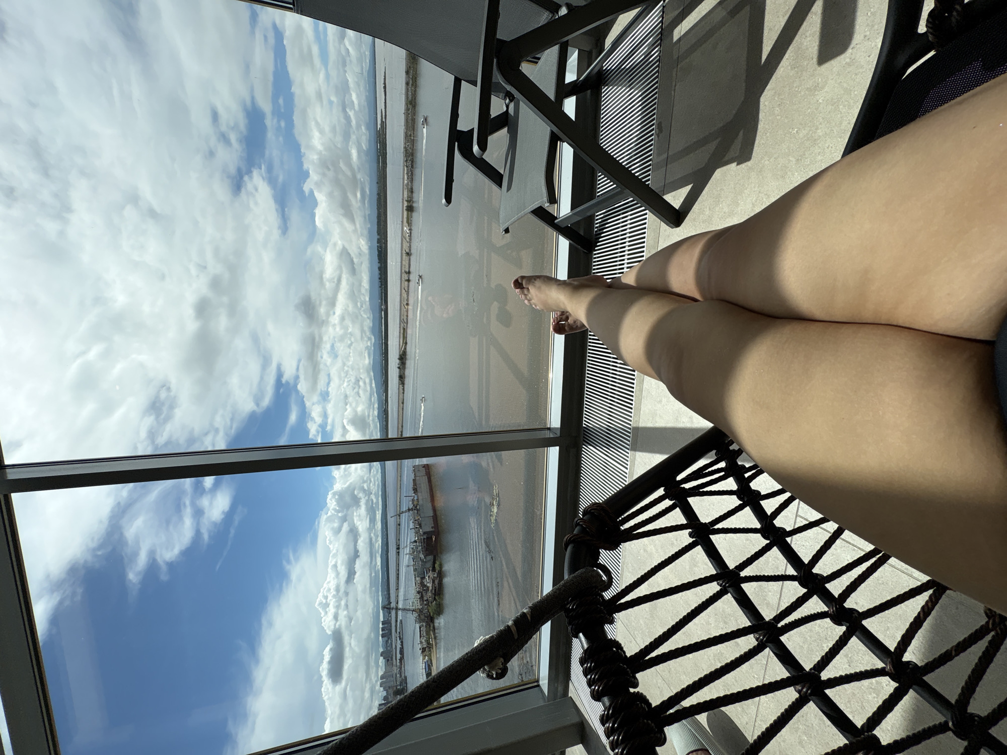 This image shows a first-person view of a person relaxing in a room with a large window overlooking a body of water and a city skyline, likely in Khabarovsk, Russia. In the foreground, a person's legs are visible, crossed, and tanned. They are sitting in what appears to be a macrame hanging chair, with the textured ropes of the chair filling the bottom left of the frame. To the right, a folding outdoor chair is positioned near the window. Through the expansive window, a wide expanse of water is visible. On the water, several boats and a large industrial structure, possibly a dry dock with cranes, are present. The background shows a distant city skyline and mountains under a cloudy sky with patches of blue. The floor appears to be tiled, and a striped ventilation grate runs along the base of the window. The overall impression is one of leisure and observation from an elevated indoor vantage point.