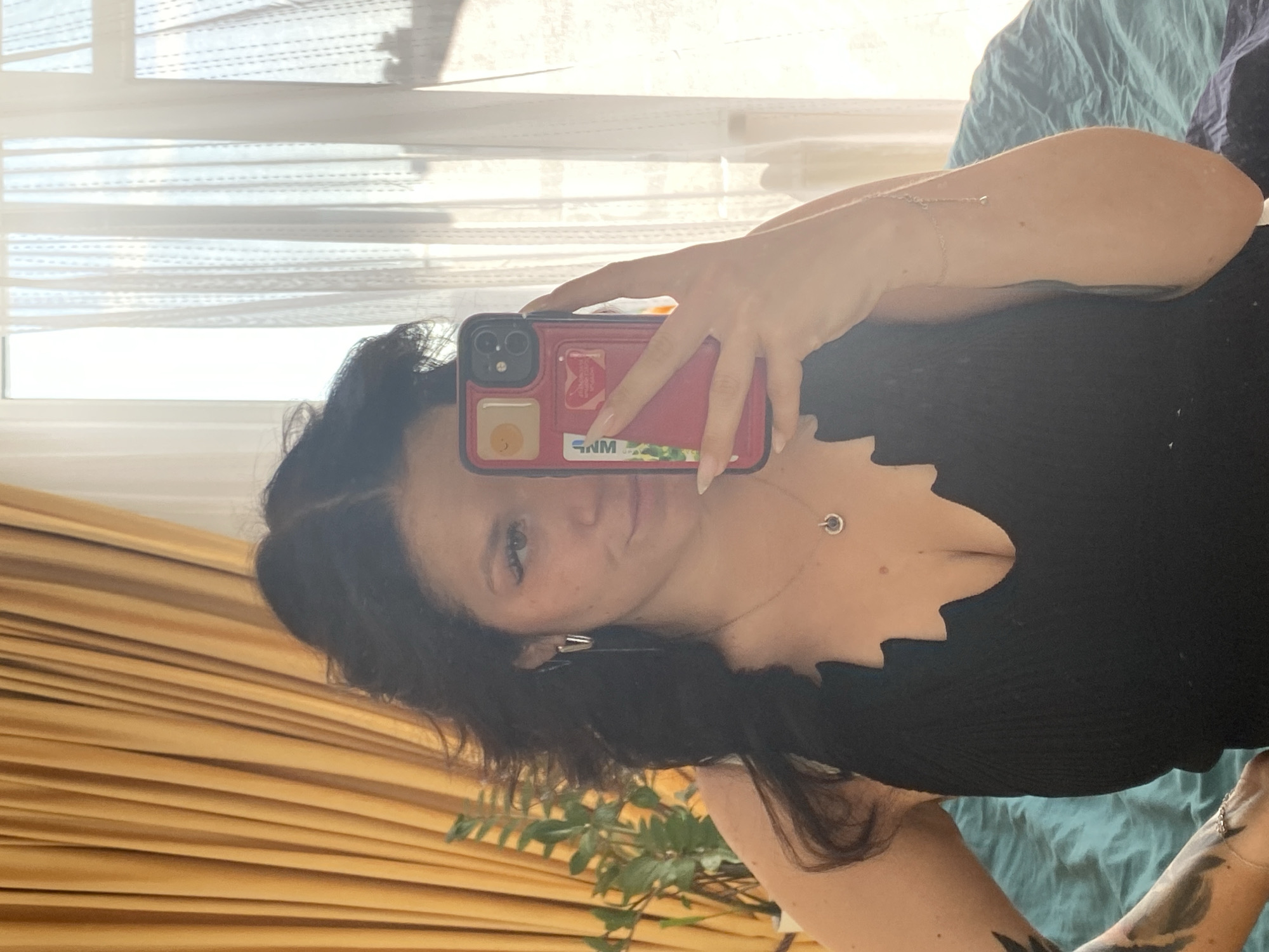 A woman with dark, wavy hair is taking a mirror selfie. She is wearing a black, low-cut top with a scalloped neckline and a silver necklace with a circular pendant. She has on hoop earrings and a delicate bracelet on her right arm. In her right hand, she holds a pink phone case with several stickers on it. Her left arm has a tattoo and is resting on her hip. She is in an indoor setting, with yellow curtains visible on the left and white sheer curtains in the background. Sunlight is streaming in from the window. The environment appears to be a bedroom, with a teal-colored bedspread visible. There is a potted plant on the left side of the image. The user's prompt is to describe the image in detail, including the location in Yanino-1, Russia. However, there is no visual information within the image that can confirm or suggest this location. Therefore, this specific detail cannot be included in the description.