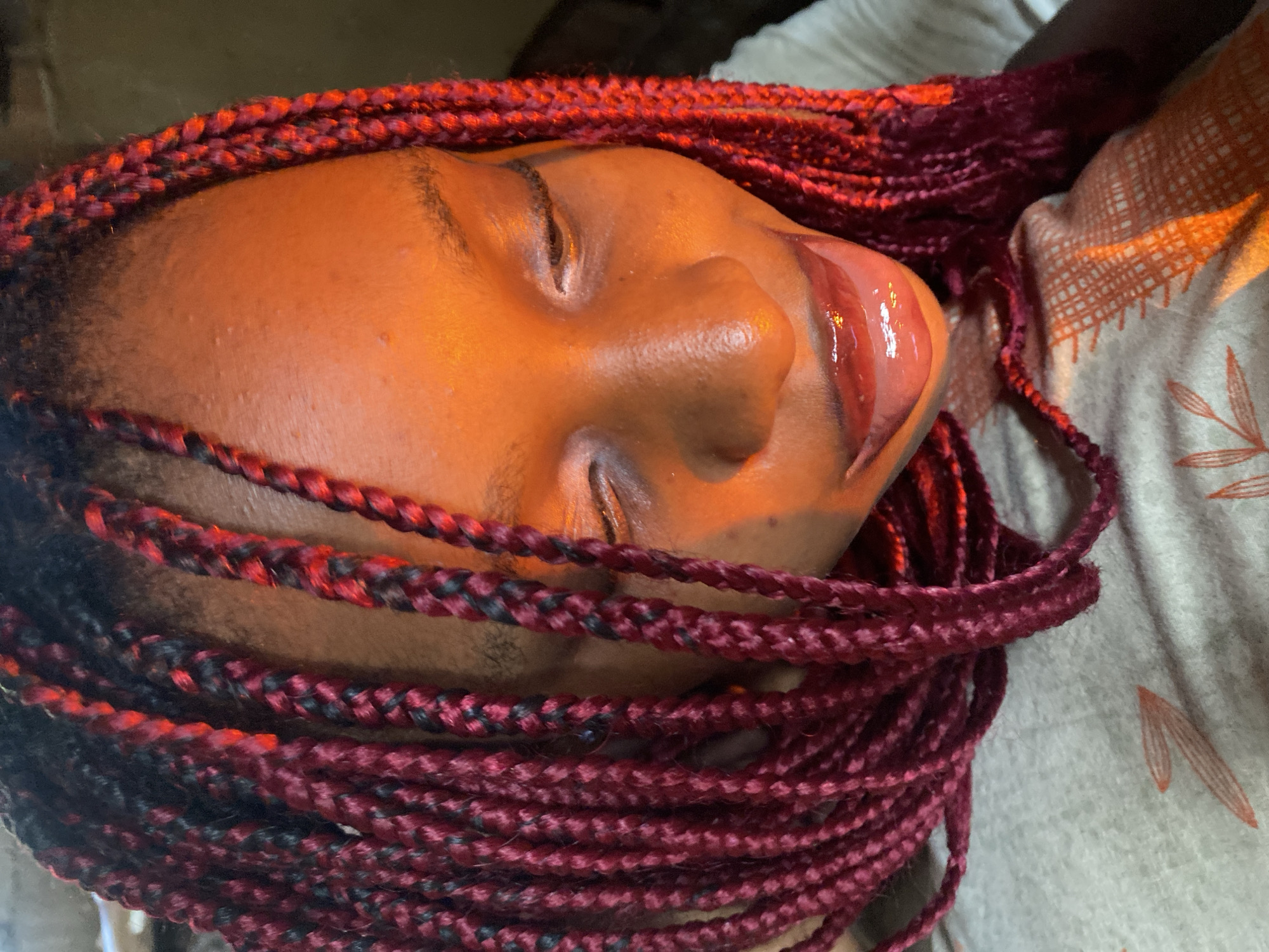 The media file is a close-up, horizontally oriented, over-the-shoulder shot of a young Black person lying down, with their eyes closed. Their face is illuminated by warm, orange light, suggesting it might be evening or a dimly lit indoor setting. The person's hair is styled in numerous small, dark red braids with some black strands, which frame their face and drape over their shoulders and the surrounding fabric.
Their lips are slightly parted, with their tongue extended and glistening, possibly from lip gloss or a similar product. The skin on their face has a warm, reddish-brown tone and appears slightly oily, with some minor blemishes visible on the forehead.
The person is lying on a textured fabric, which is light gray with a pattern of thin, orange, leaf-like shapes. The overall impression is one of relaxation or being at rest. There is no visible text in the media file. The setting is implied to be Iranla, Nigeria, based on the provided location context.