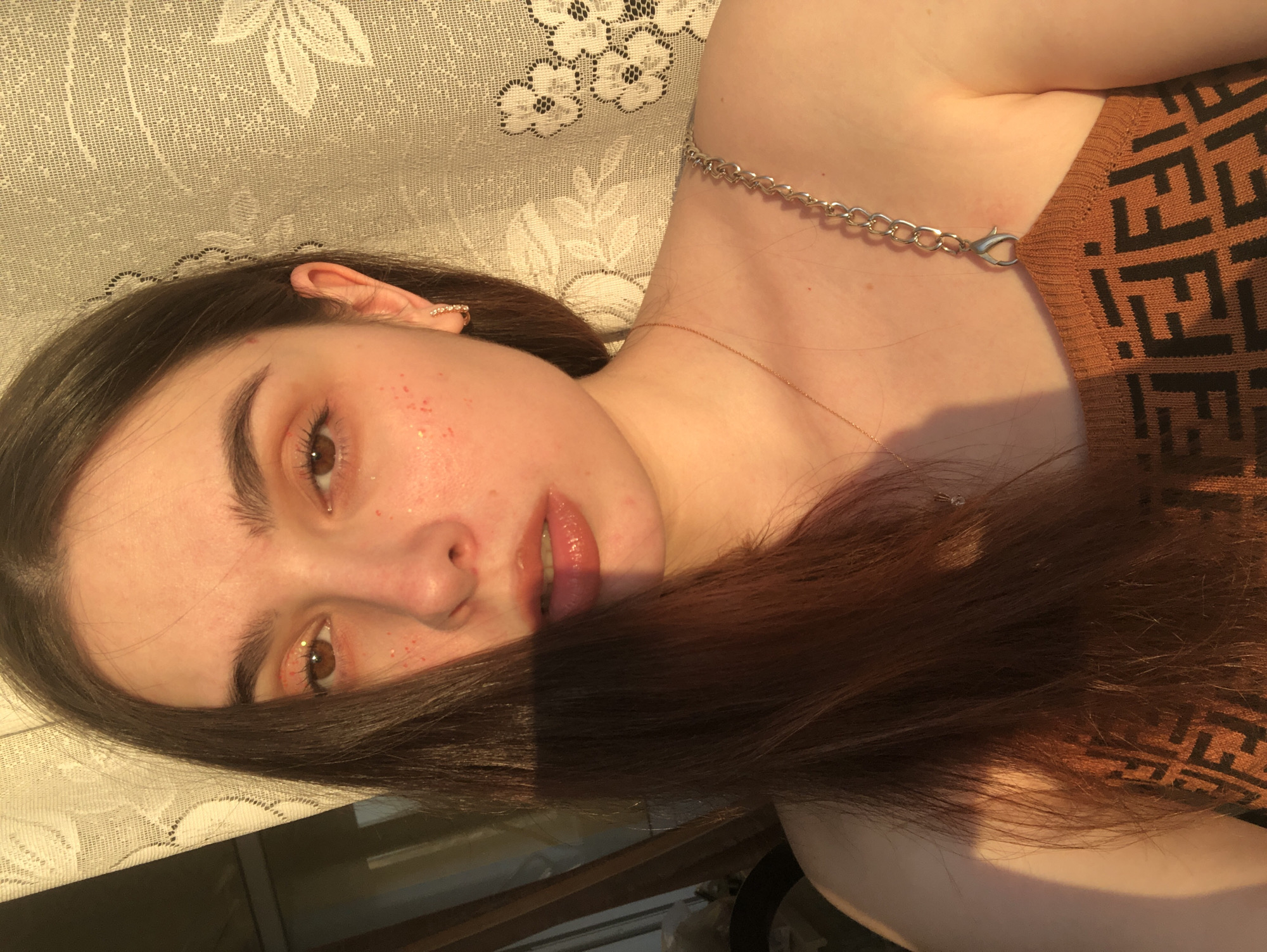 A young woman is lying on a surface covered with a lace-patterned textile. She is partially facing the camera, with some of her brown hair obscuring part of her face. She has fair skin with some visible freckles around her eyes and nose. She wears a brown top with a black pattern, and a silver chain necklace. Part of the bottom of an undefined object is visible in the lower left corner of the image. The light suggests an indoor setting during daylight hours. No specific location in the city of Noviy, Russia is discernable from the image.