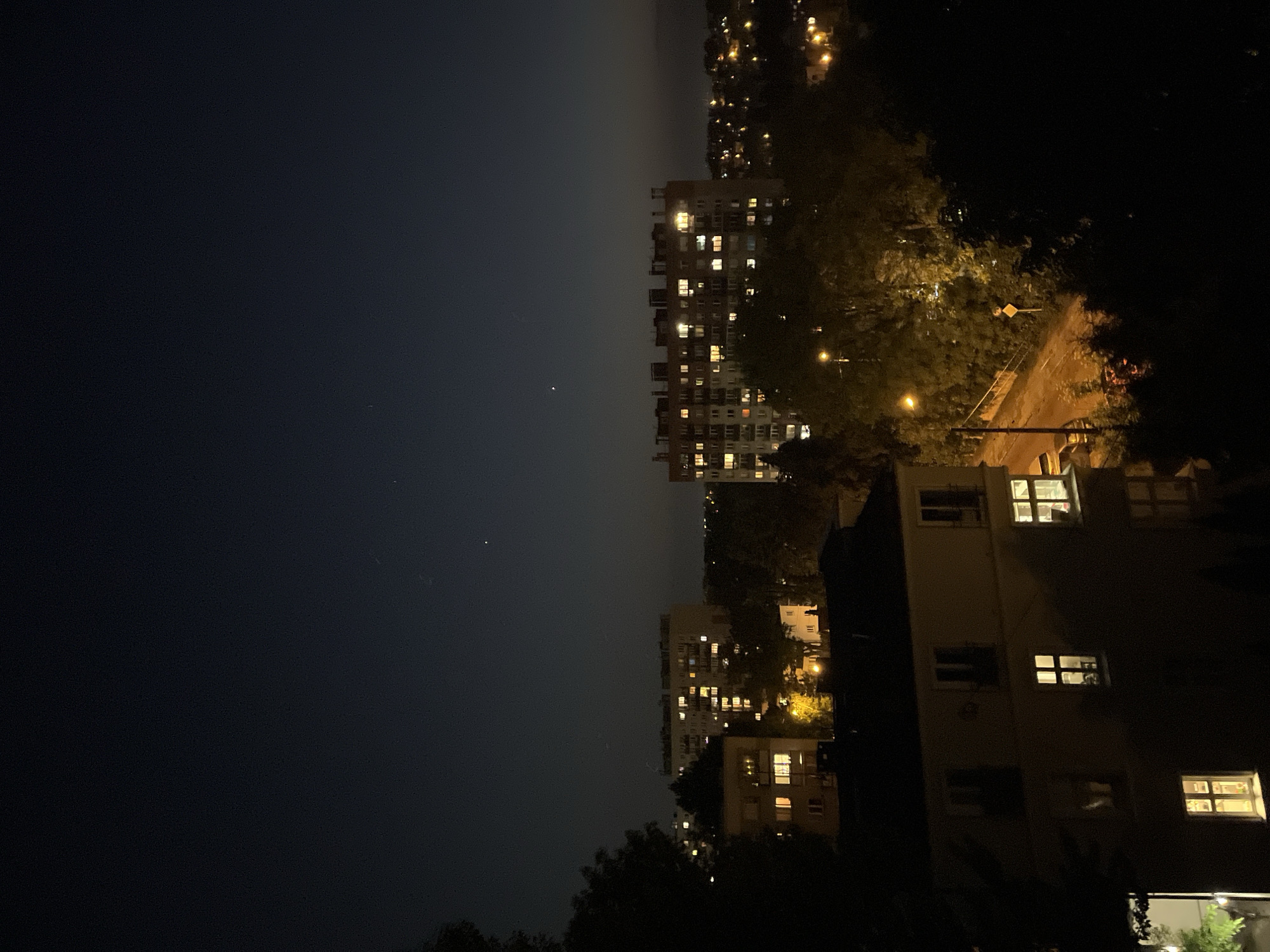 The image shows an urban night scene in Unknown, Unknown. On the left is a dark sky. In the center are tall apartment buildings with lit windows. To the right is foliage and another building. A road and street lights are also visible.