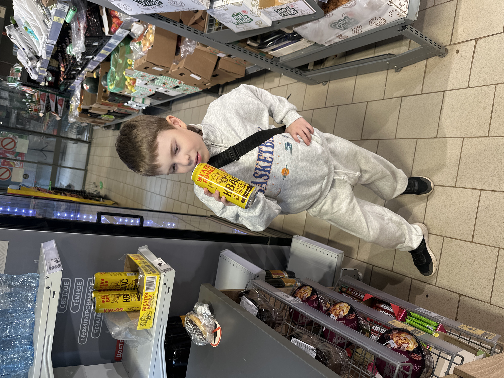 A young boy stands in a grocery store aisle, holding a yellow can of "Zhelataya Bochka" kvass. He is wearing a light gray tracksuit and black sneakers. The store shelves are stocked with various food items, including bottled water, canned drinks, and packaged goods. Some shelves display signs with Cyrillic text such as "Svetloe," "Tyomnoe," and "Nefiltrovannoe." The boy appears focused on the can he is holding. The lighting suggests it is daytime, and the setting is indoors. The overall scene depicts a typical shopping experience in a Russian grocery store.