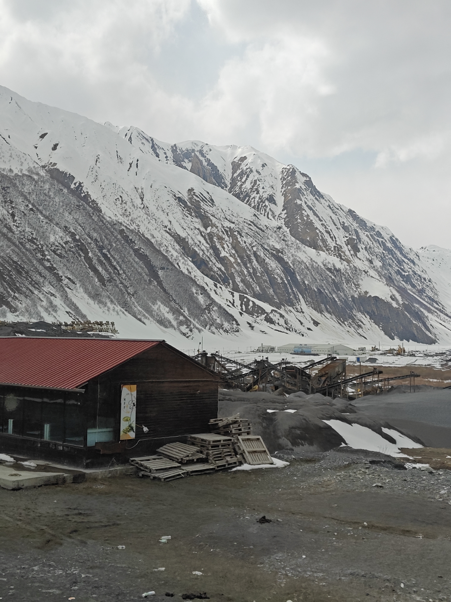 The image displays a wide landscape dominated by towering, snow-capped mountains under an overcast sky. The mountains in the background are extensively covered in white snow, with darker patches of exposed rock and sparse vegetation visible on their slopes.
In the left foreground, a rustic-looking building with dark wooden walls and a corrugated red metal roof stands prominently. This building features large glass windows, and a yellow banner or poster is affixed to its side, displaying the letters "MFF" and images of various fruits, including lemons, limes, and grapes. Nearby, a large stack of wooden pallets is visible on the ground.
To the right of this building and stretching into the mid-ground is an extensive industrial operation. This complex includes numerous grey conveyor belts, crushers, and sorting equipment, indicating a gravel or aggregate processing plant. Large piles of dark grey, crushed material are accumulated around the machinery, with patches of snow interspersed. Further into the distance, at the base of the mountains, several white industrial buildings and what appears to be a yellow excavator can be faintly seen.
The ground in the foreground is a mix of gravel, dirt, and muddy patches, with scattered plastic bottles and other litter. The overall weather is cold, as evidenced by the widespread snow and the grey, cloudy sky. No people are visible in the scene, but the presence of industrial machinery suggests ongoing or recent operations. The time of day appears to be daytime, though the heavy cloud cover creates a muted, uniform light.