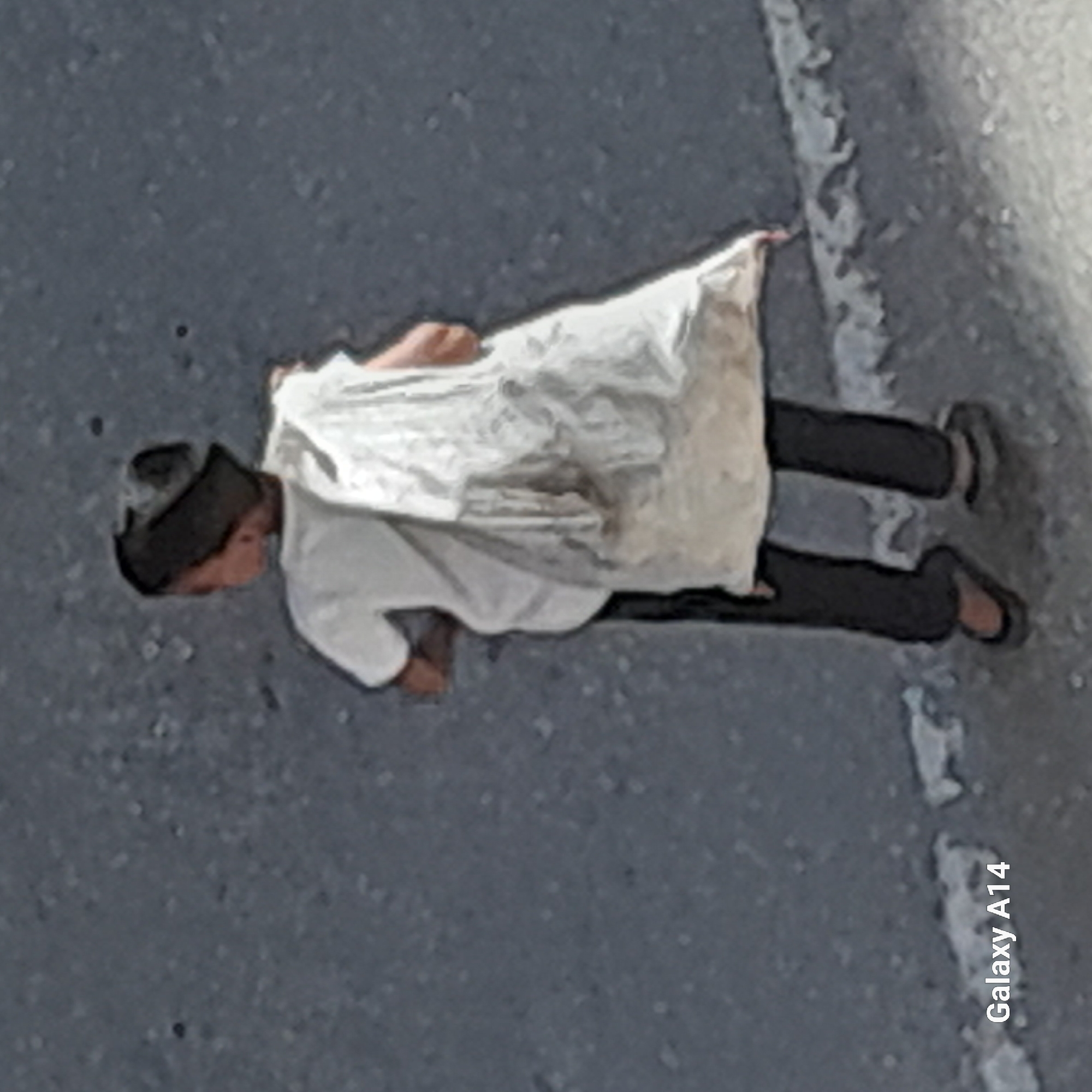 The image captures a single person, likely a child or young male, engaged in a physical activity resembling a push-up on a dark, paved outdoor surface. The person is wearing a dark cap, a loose-fitting white long-sleeved garment (possibly a thobe or tunic) over a white shirt, dark trousers, and dark sandals or slip-on shoes. Their hands are placed on the ground, supporting their upper body, with their head slightly lowered.
The setting appears to be an outdoor area with a dark grey, speckled ground, consistent with asphalt or concrete. A faded and somewhat uneven white line runs parallel to the person's body on the paved surface. The general lighting suggests it is daytime.
The image itself is rotated 90 degrees clockwise and is of low resolution and appears grainy, which is also hinted at by the visible watermark "Galaxy A14" in white text at the bottom right corner, indicating the camera used to capture the photo. No specific location details or discernible emotions are present.
