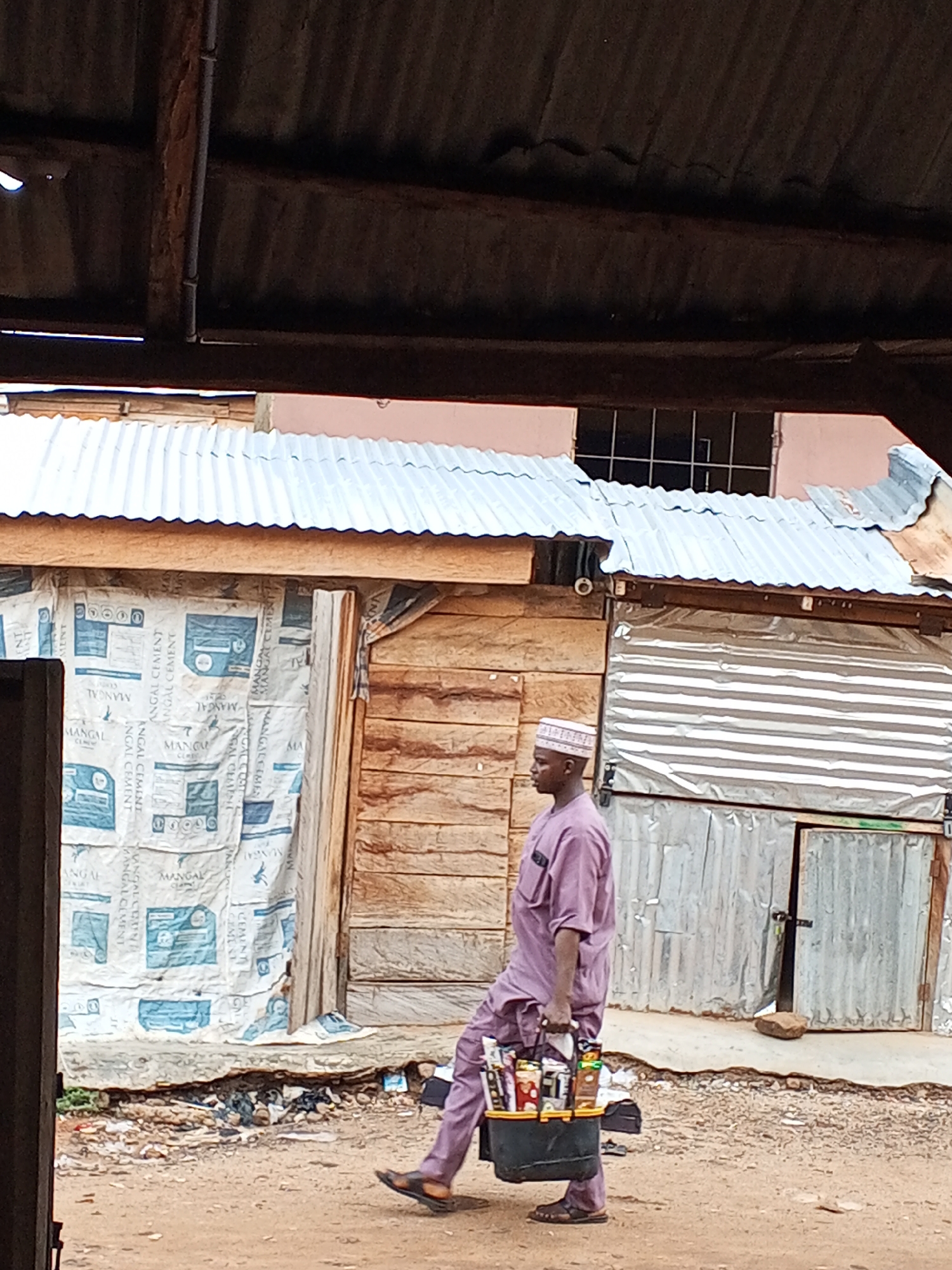A man is walking in Lokoja, Nigeria, carrying a black bin filled with what appears to be snacks and drinks. He is wearing a light purple outfit and a small white cap, and he is wearing sandals. The surroundings suggest a relatively impoverished area. The buildings appear to be made of wood, corrugated metal, and cement block. There is a banner advertising Mangal cement. The ground is dirt. The scene is framed by part of a roof.