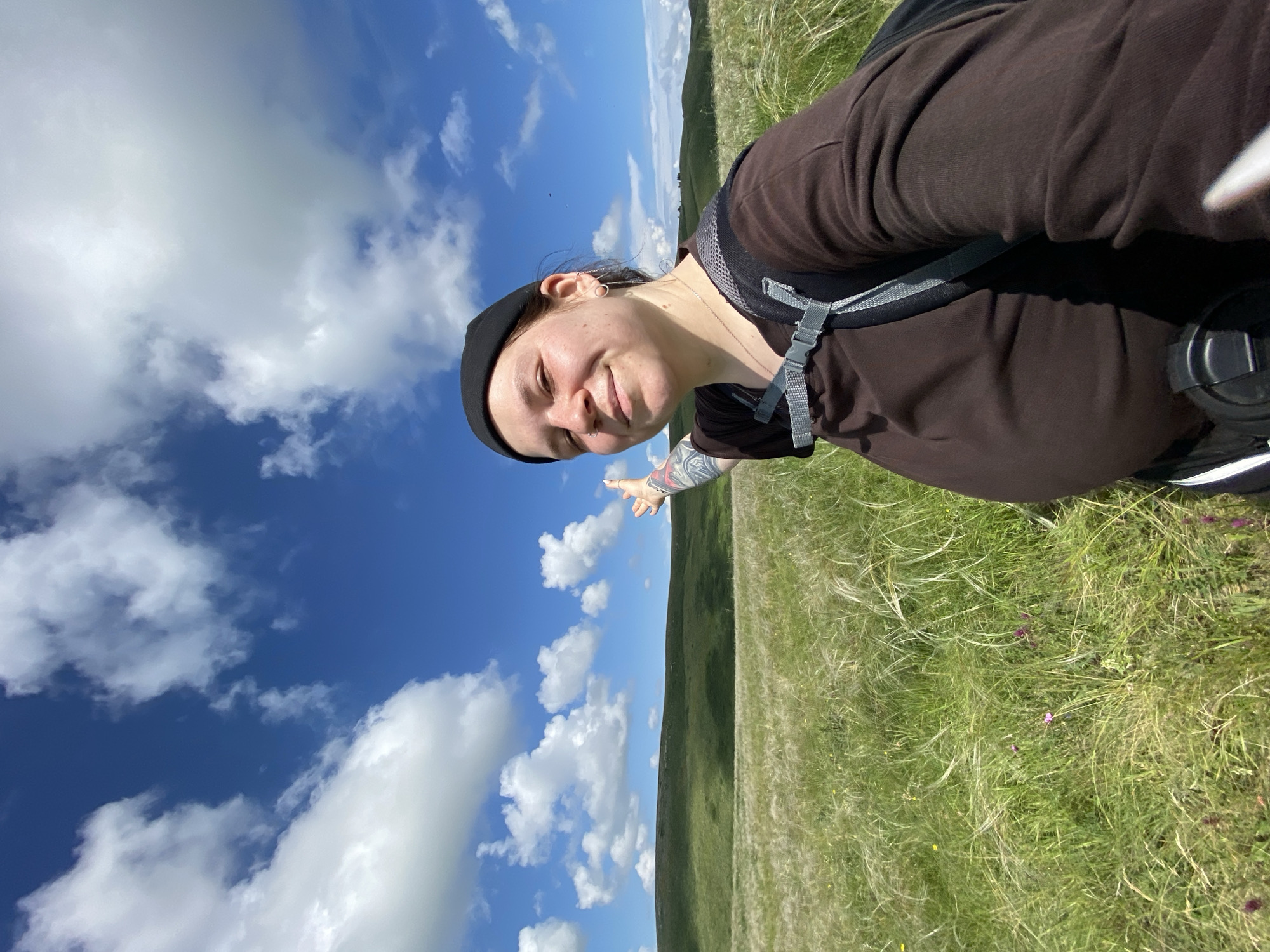 A young person, wearing a black beanie, a brown long-sleeved shirt, and a backpack, is taking a selfie in a grassy, hilly landscape under a bright blue sky with scattered white clouds. The person's left arm is raised and pointing towards the sky. The background shows rolling green hills and a vast expanse of dry grass, characteristic of the natural landscape around Bogoslovka, Russia. The image captures a sunny day, likely during a hike or outdoor excursion.