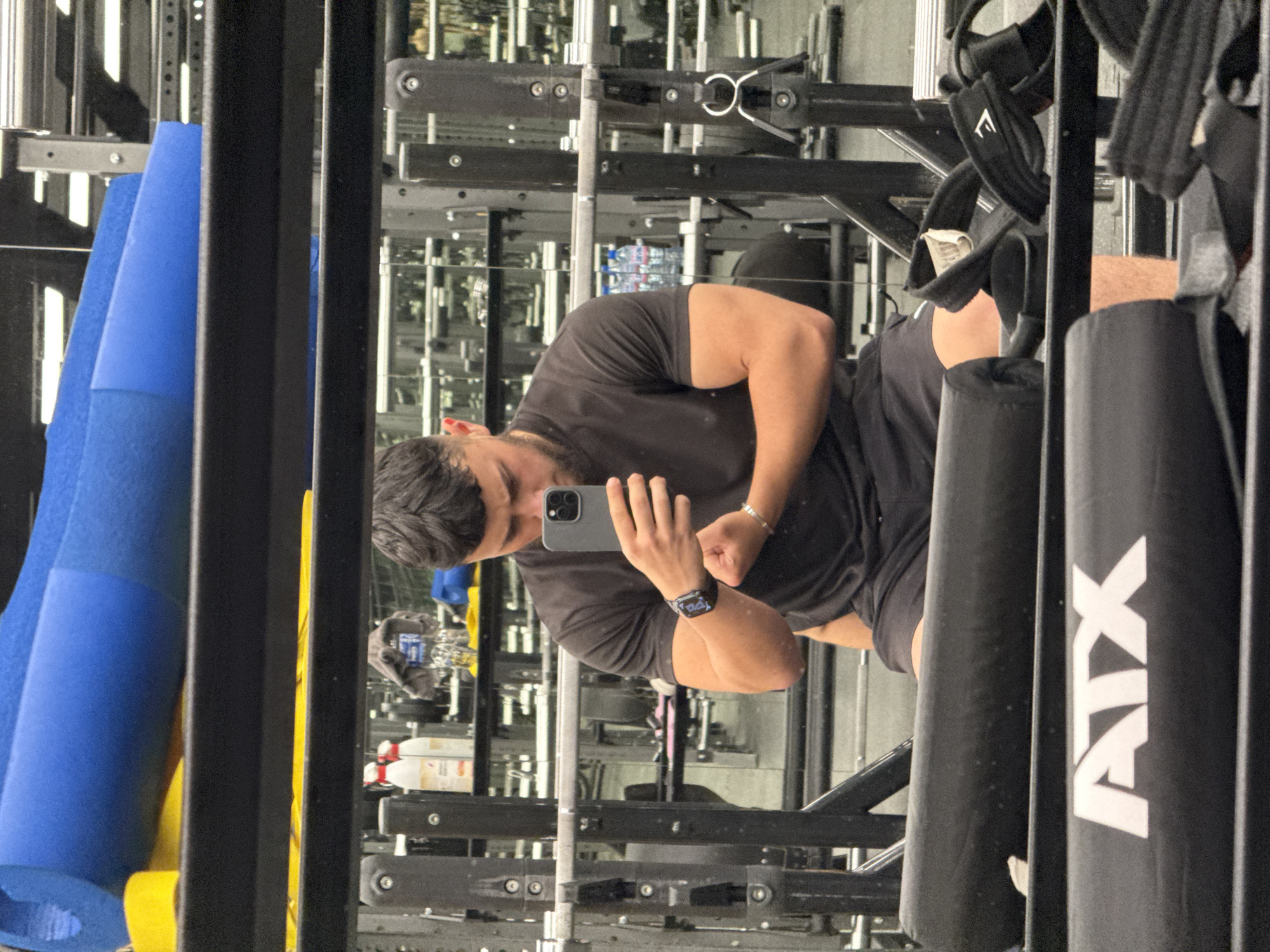 A young man is taking a mirror selfie in a gym. He is wearing a black t-shirt and shorts. He is flexing his bicep and looking at his phone. The gym appears to be well-equipped with weightlifting machines and free weights. There are also some yoga mats in the background. The scene is well-lit and the overall atmosphere suggests a serious workout session.