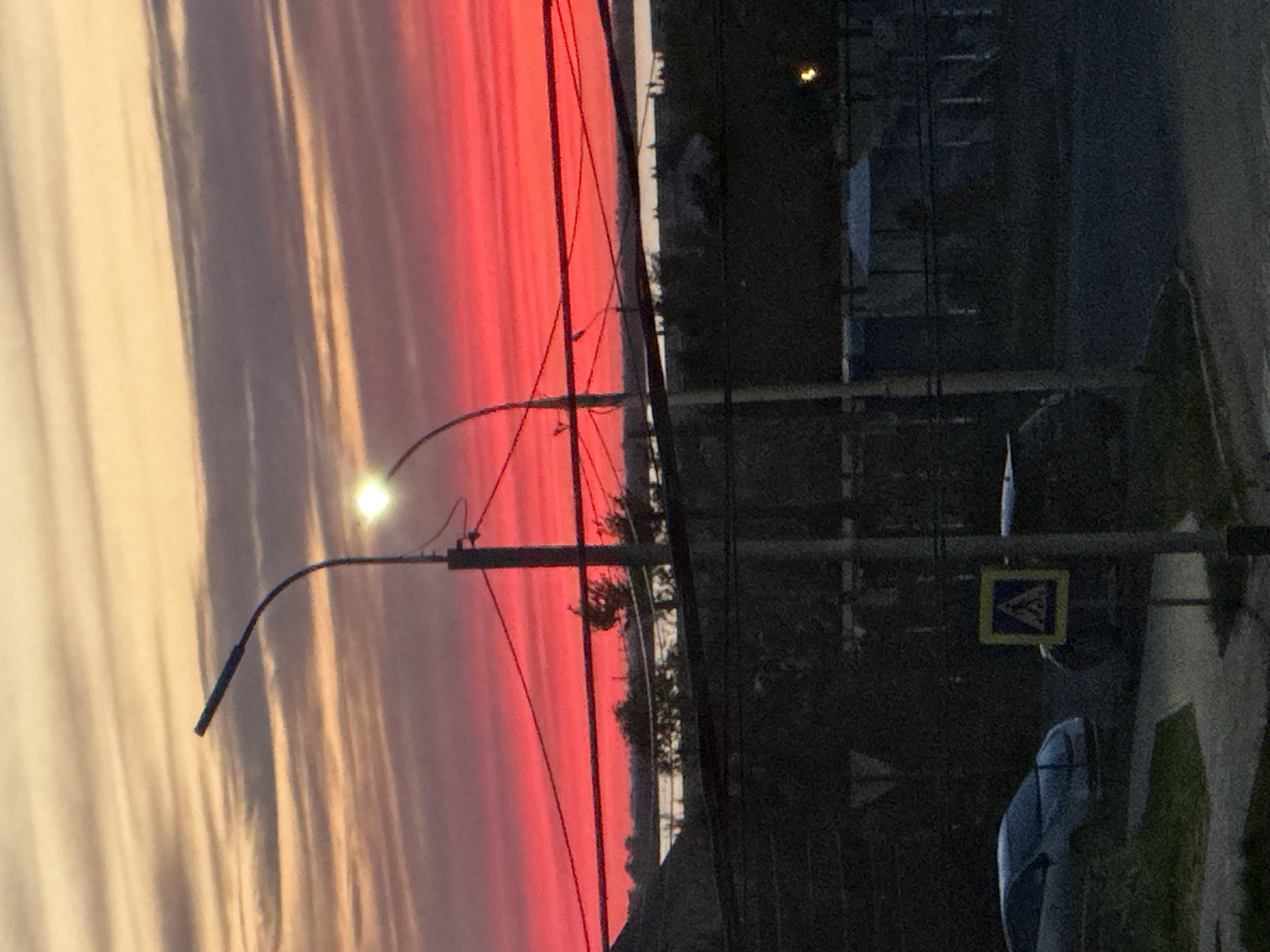 The image captures a street scene at dusk or dawn in what is possibly Khabarovsk, Russia, based on the architectural style and street signs. A vibrant red and orange sky dominates the left half of the frame, indicative of a sunrise or sunset. On the right side, there's a street scene. The immediate foreground includes a section of sidewalk or curb, and a car. There are buildings or structures behind the street, obscured by what seems to be power lines and streetlights. The scene is illuminated by a street lamp, the source of a strong white light. The overall impression is of a quiet urban scene under a dramatic sky. The photo appears to be taken from a ground-level perspective, possibly by a handheld camera.