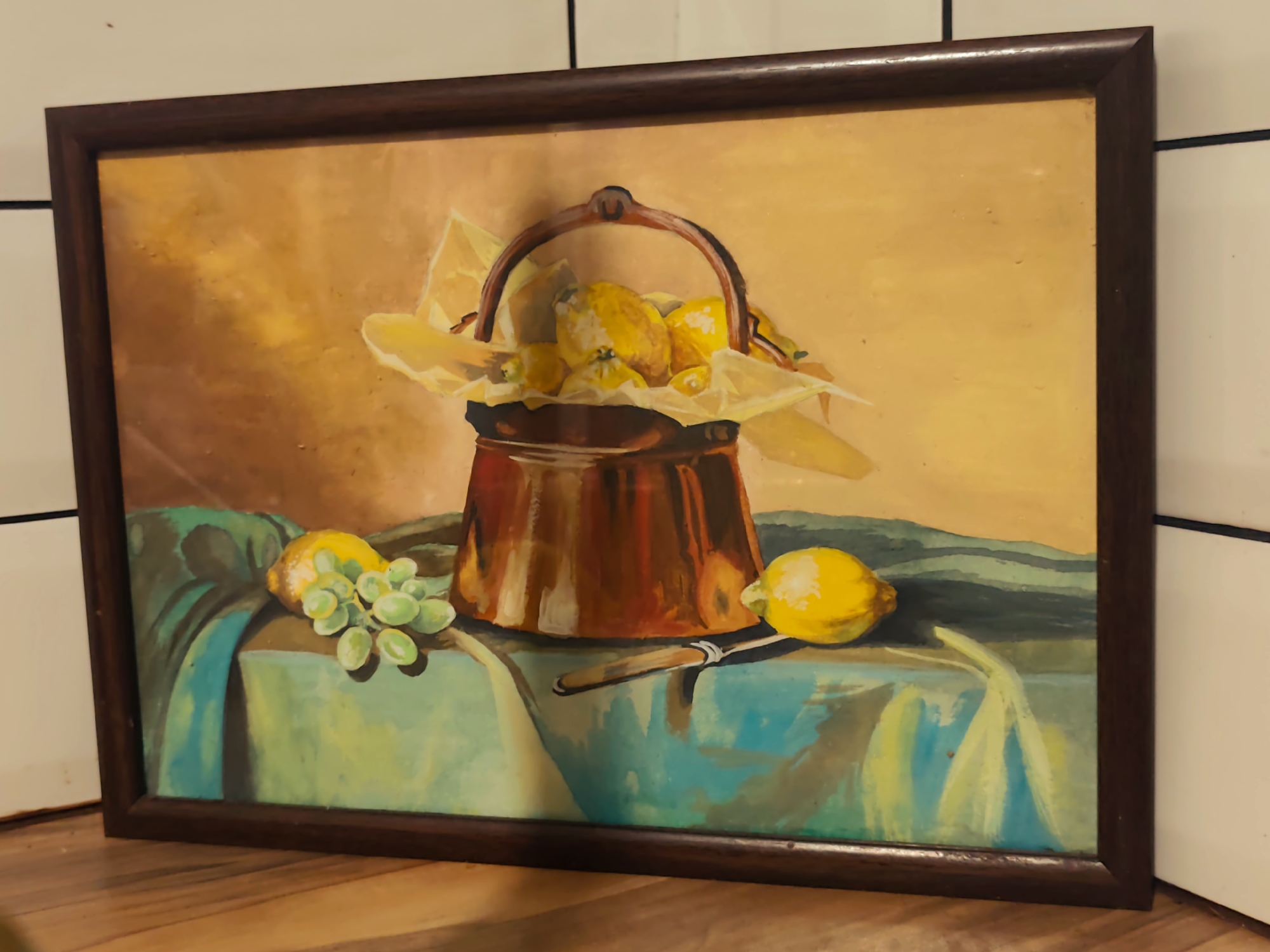 A framed still life oil painting depicts a copper pot filled with lemons and paper, placed on a teal tablecloth. To the left of the pot are a bunch of green grapes and a single yellow lemon. A knife with a light-colored handle rests on the tablecloth near the pot. The background is a warm yellow, and the painting is framed in dark wood. The painting is positioned on a wooden surface, against a tiled wall.